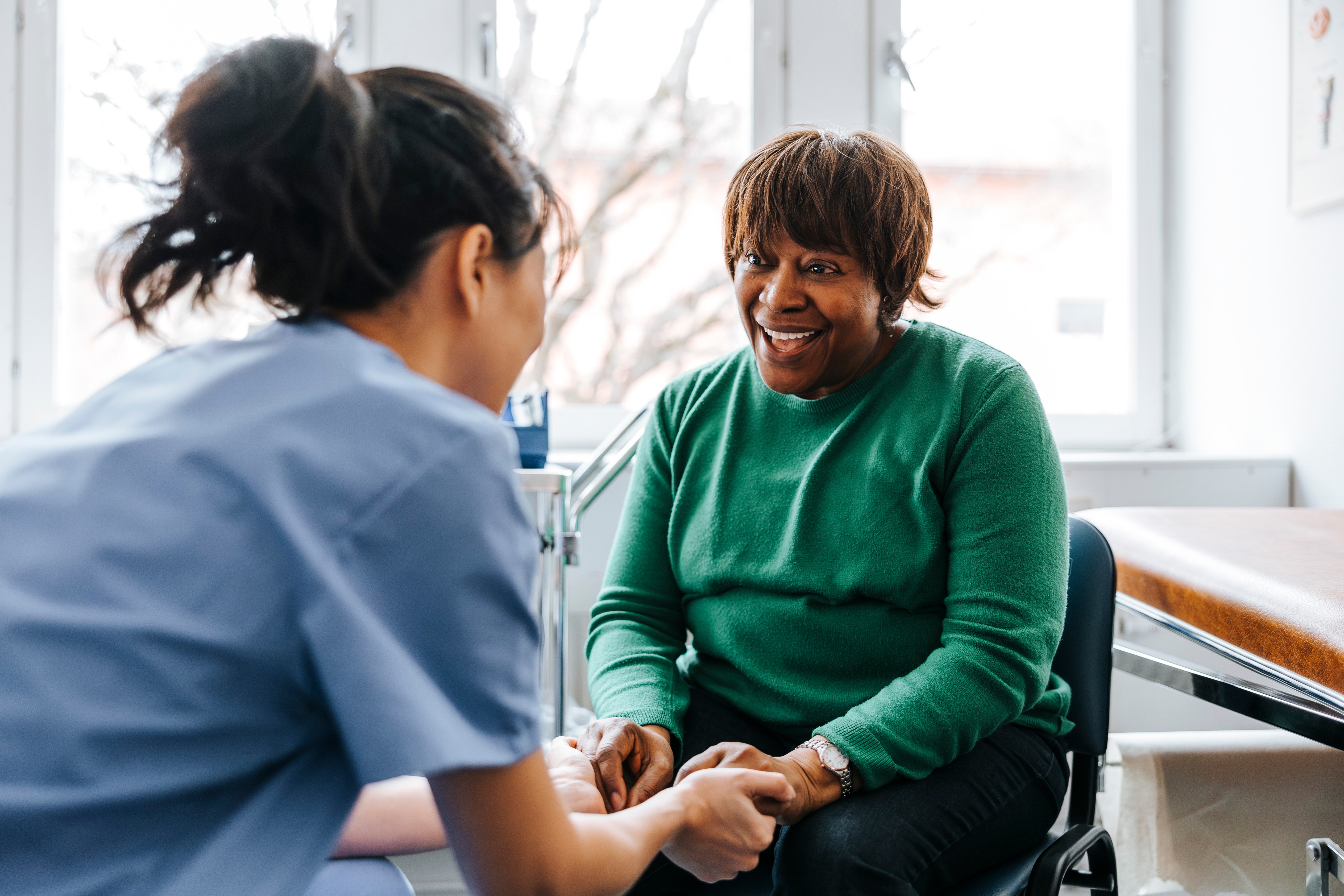 En sykepleier i scrubs rådfører seg med en smilende kvinne som sitter på et medisinsk kontor, holder hendene hennes og gir trøst