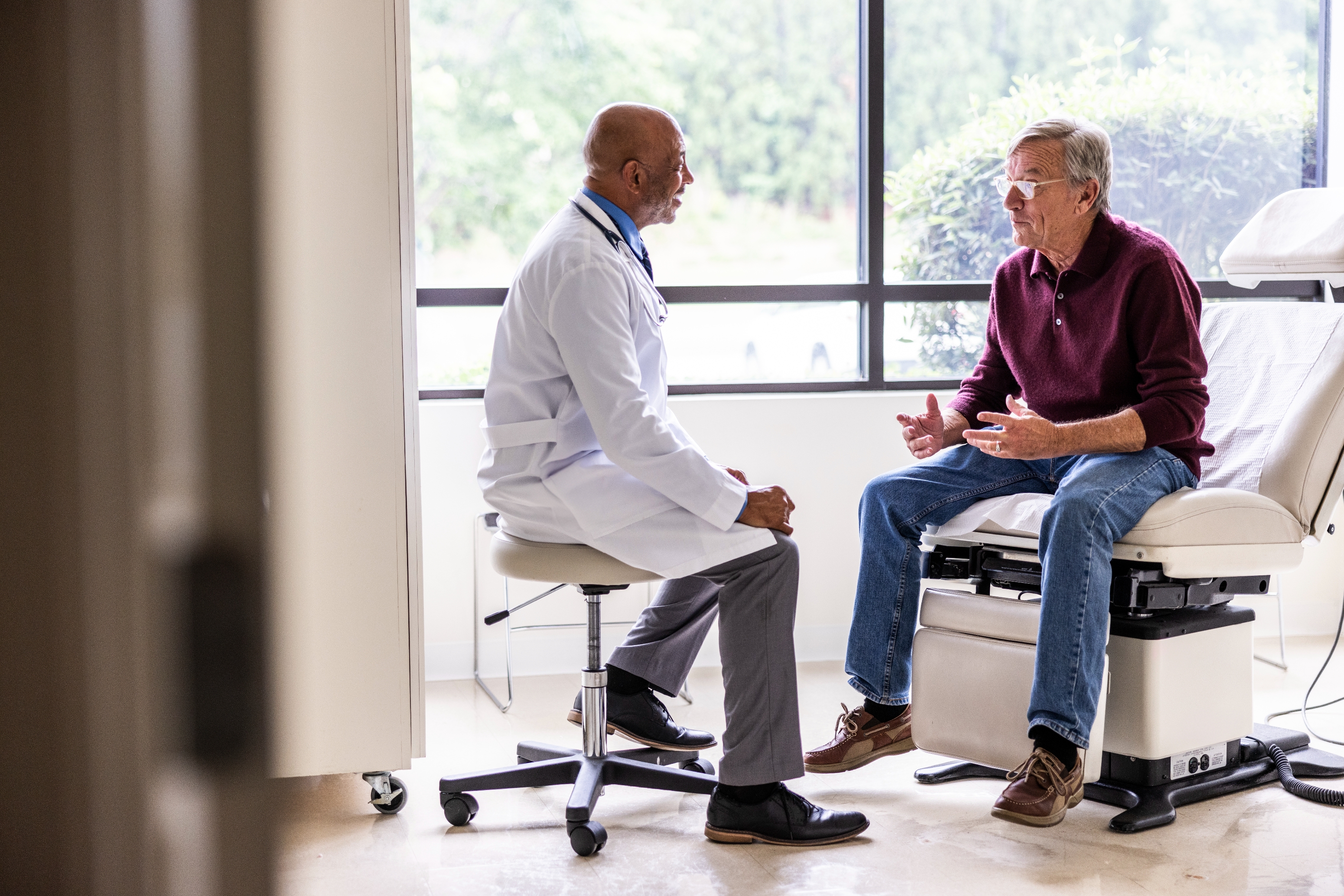 Lege og eldre voksen mann på et konsultasjonsrom, sittende og engasjert i en alvorlig samtale ved vinduet