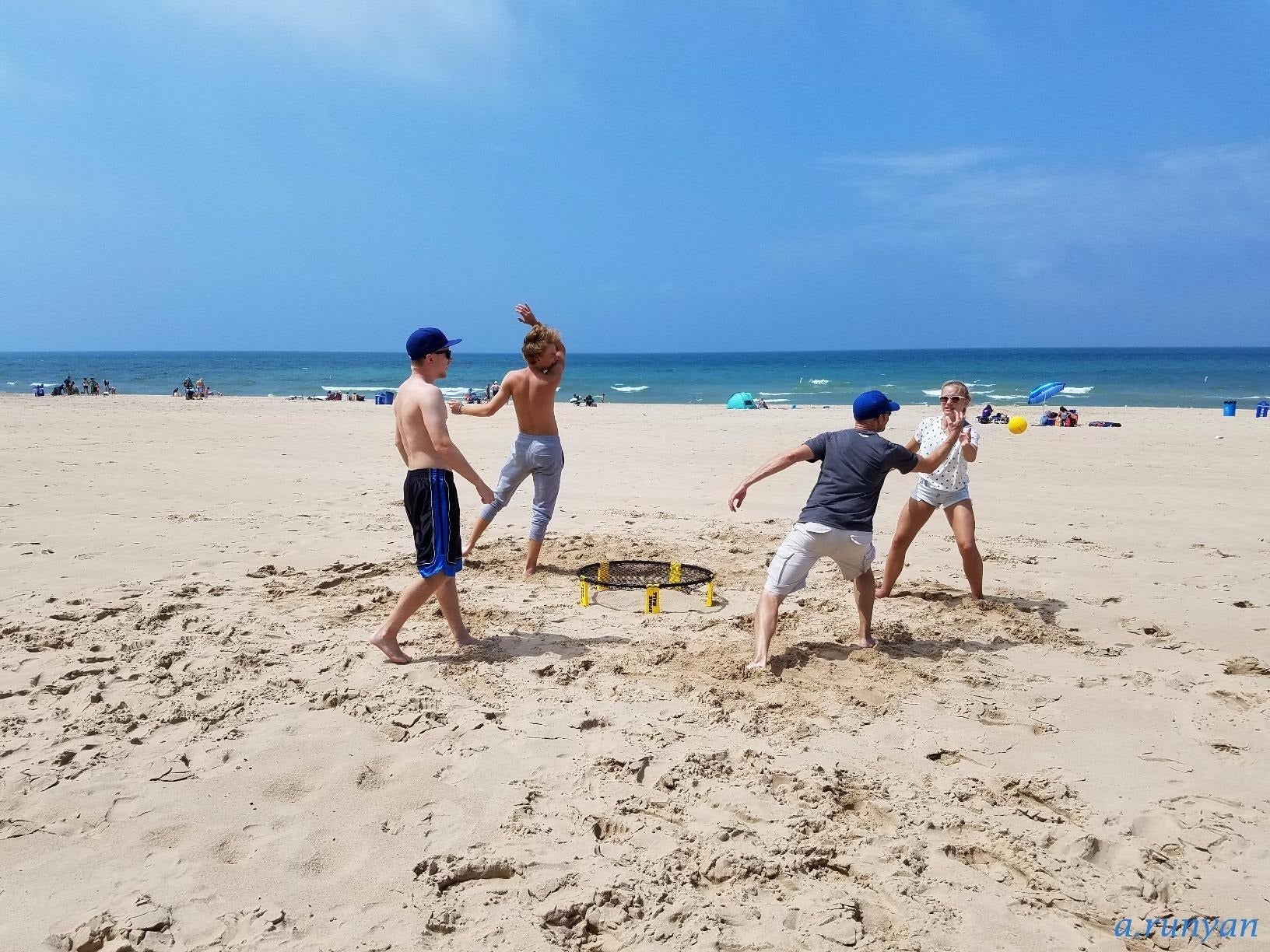Four people play beach Spikeball, jumping and diving in the sand near the ocean