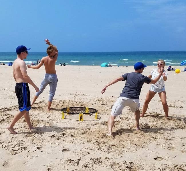 Four people play beach Spikeball, jumping and diving in the sand near the ocean