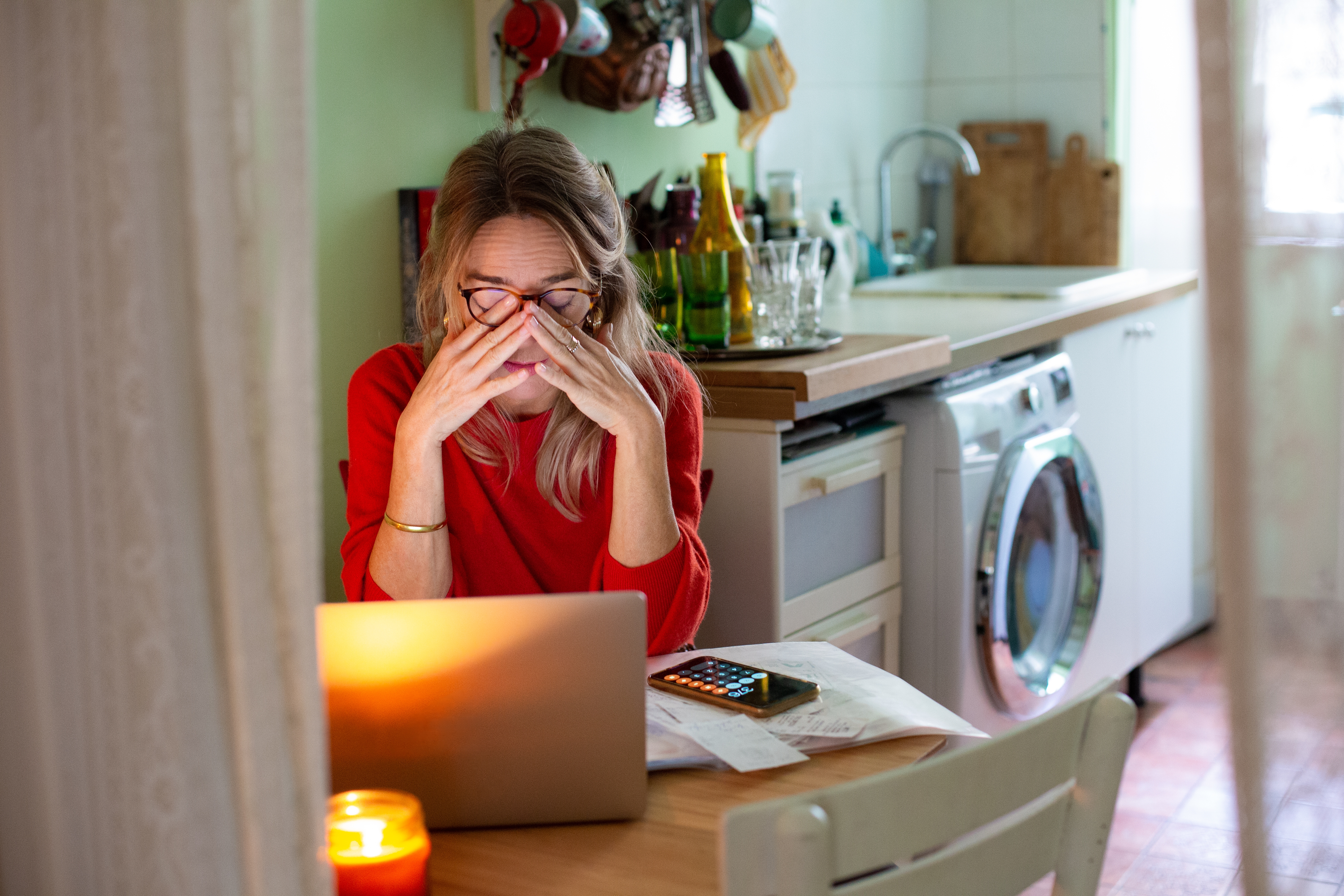 En person på et kjøkken ser stresset ut mens han bruker en bærbar datamaskin, omgitt av papirer og en kalkulator