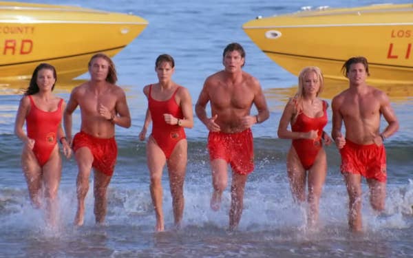 Six lifeguards in red swimsuits run out of the ocean, with yellow rescue boats in the background, in the baywatch intro