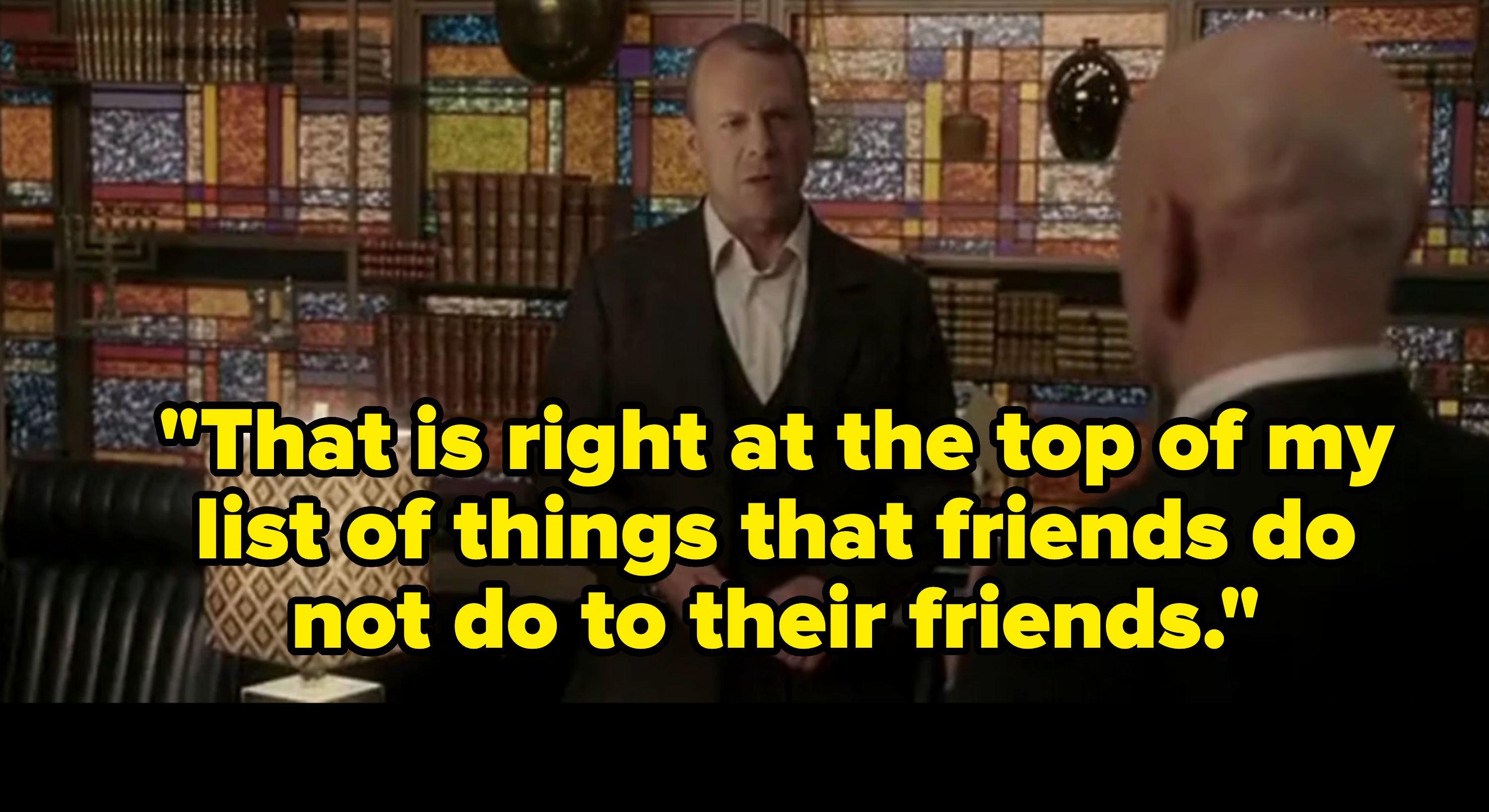 Two people in a study-like room with a stained glass wall and books. One person is wearing a suit