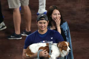 野球選手がユニフォーム姿で犬を抱え、楽しそうに歩いている。後ろにカジュアルな服装の人が続く。