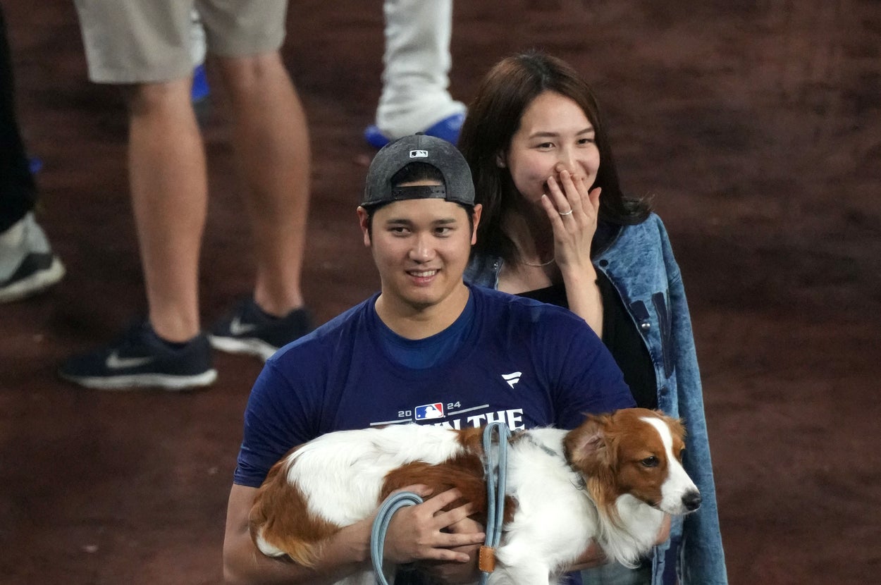 野球選手がユニフォーム姿で犬を抱え、楽しそうに歩いている。後ろにカジュアルな服装の人が続く。