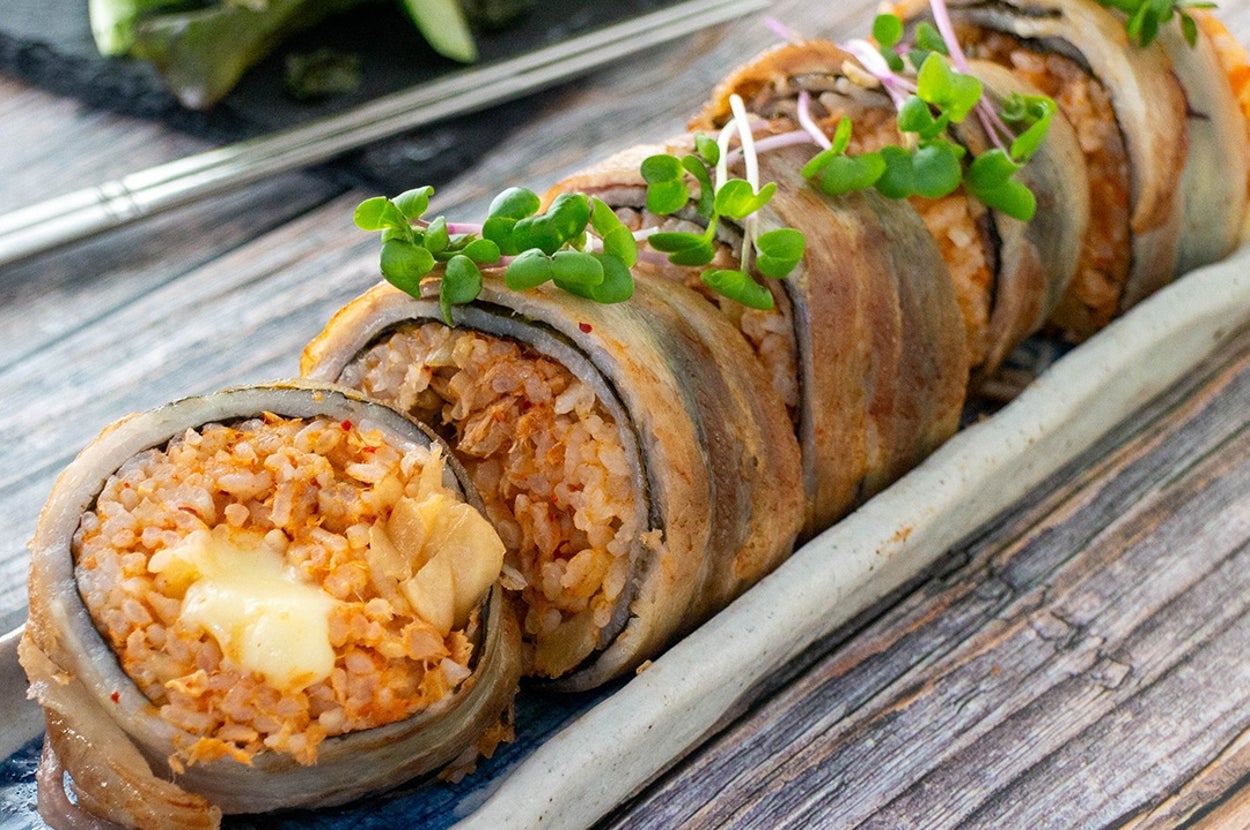 木製テーブルの上にある陶器の皿に盛られたベーコン巻きのキムチ焼き飯ロール。隣に緑のサラダが添えられている。