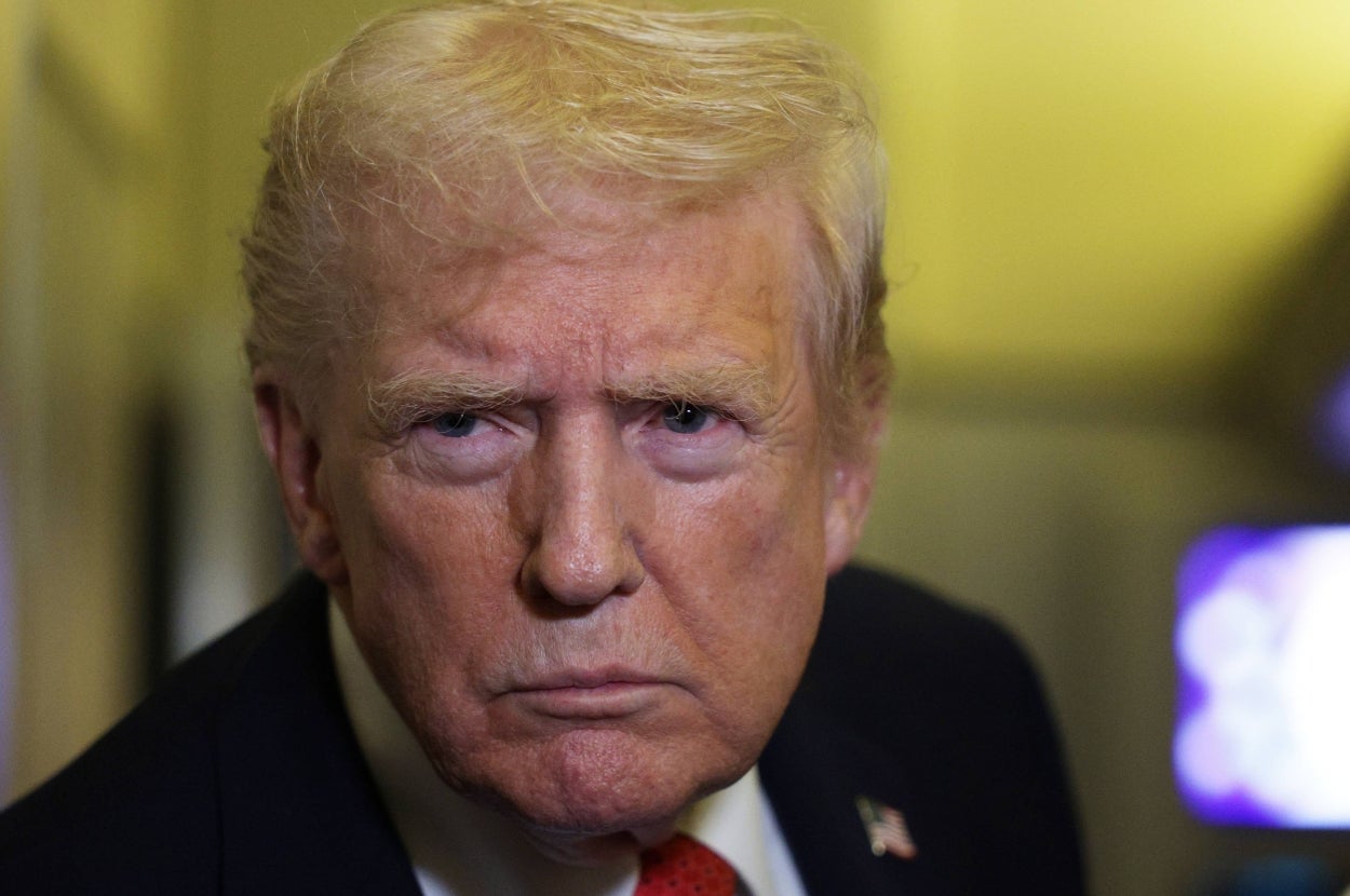 A person with a serious expression, wearing a suit, appears to be at a formal or media event. Background is blurred
