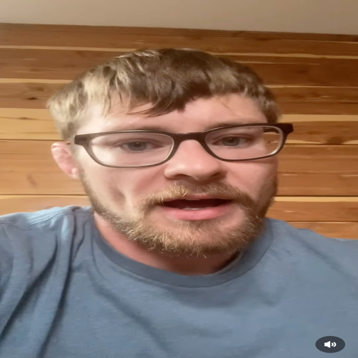 A person with glasses and a beard speaks directly to the camera, in a room with wooden paneled walls