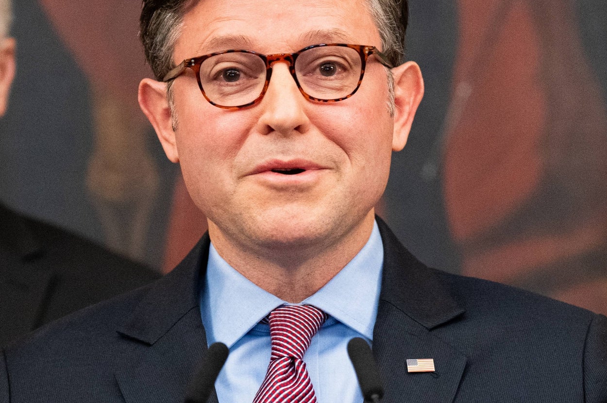 Man in glasses and suit speaking at a podium with microphones