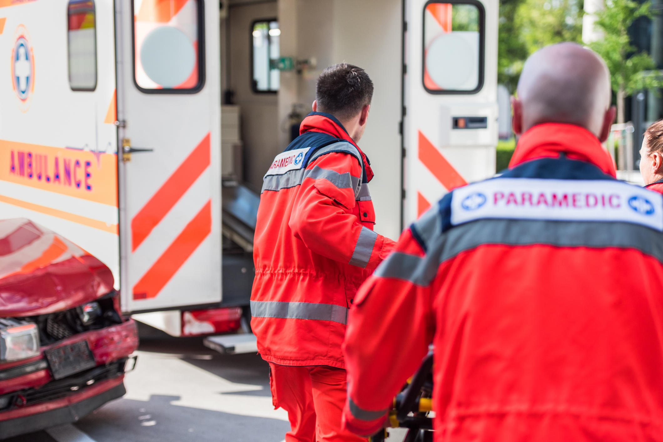 Ambulansepersonell i uniform bistår med en båre ved siden av en ambulanse og skadet kjøretøy på et gatebilde