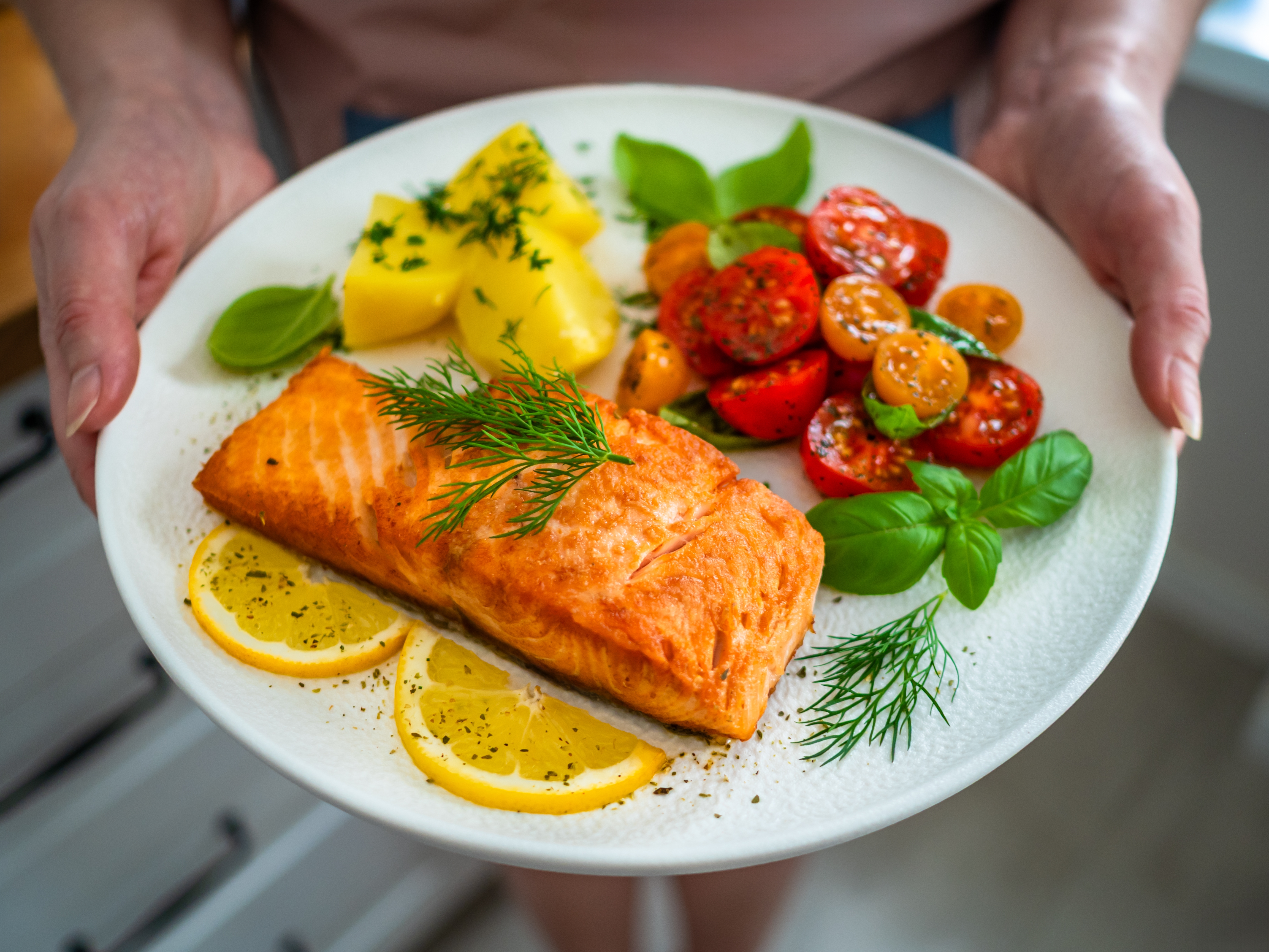 Person som holder en tallerken med grillet laks, poteter, cherrytomater, fersk basilikum og sitronskiver, klar til servering