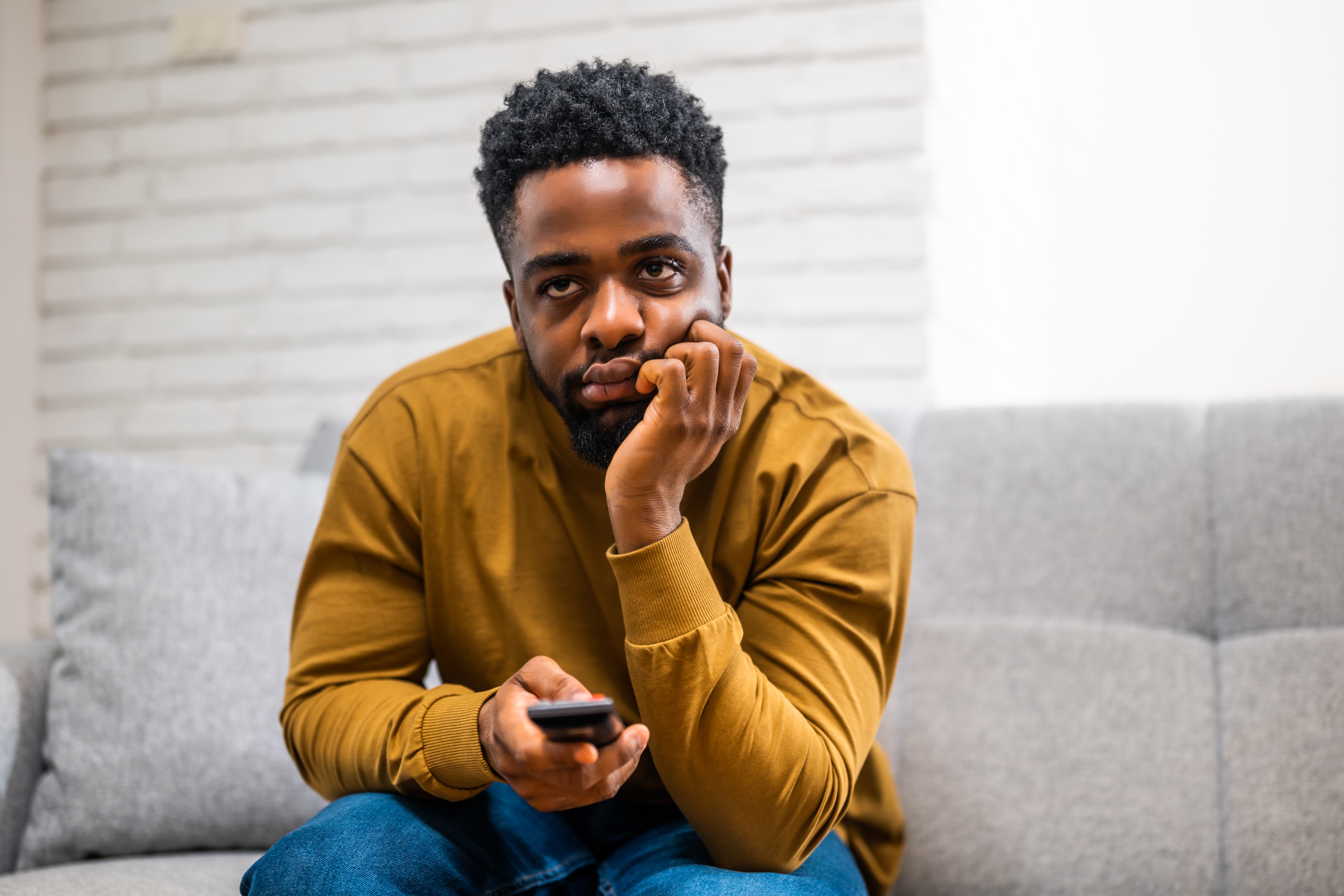 Person i uformelle klær som sitter på en sofa, holder en fjernkontroll, ser ettertenksomt på noe