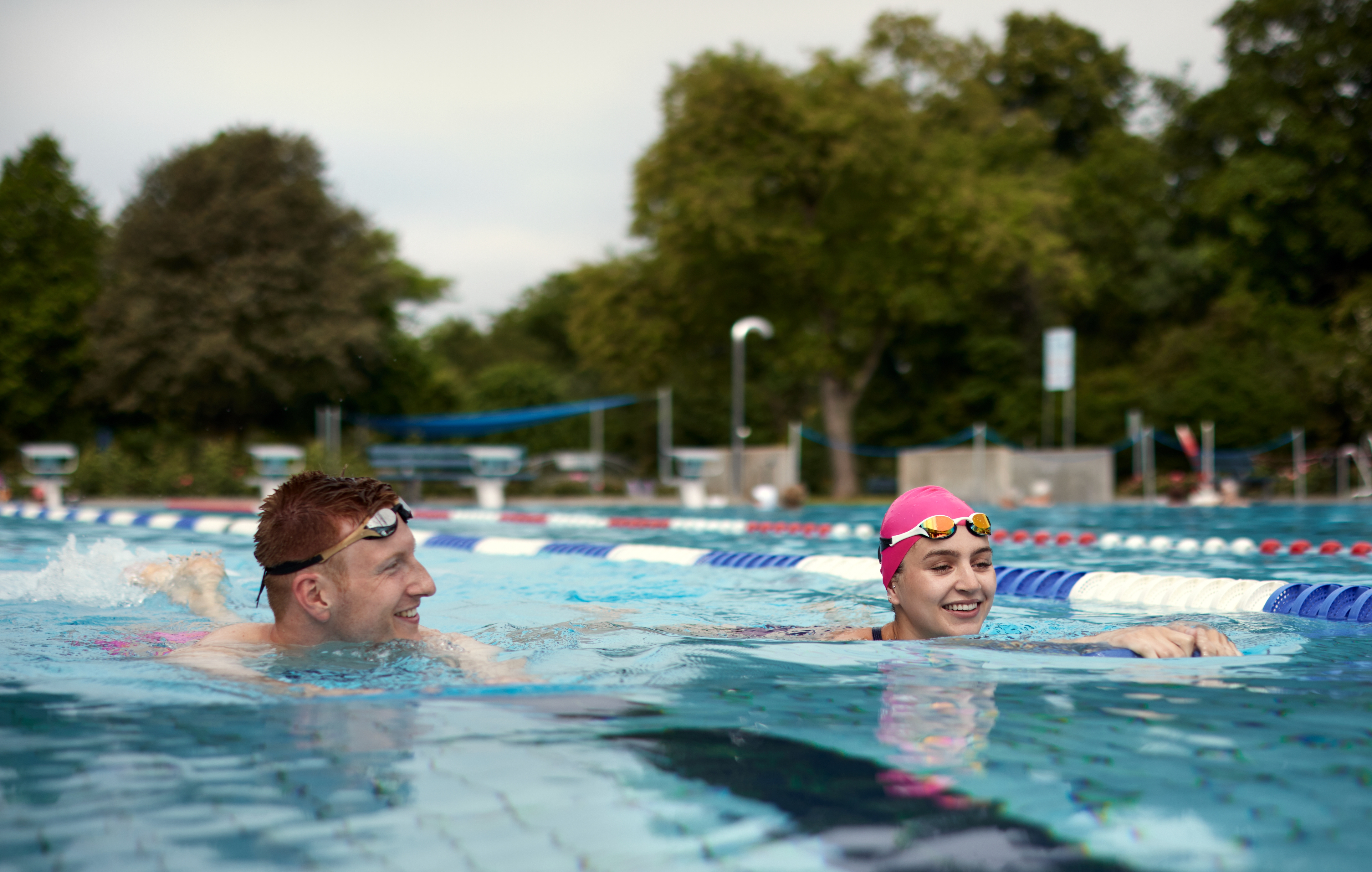 To personer svømmer i et utendørsbasseng, iført svømmehetter og briller, med trær i bakgrunnen