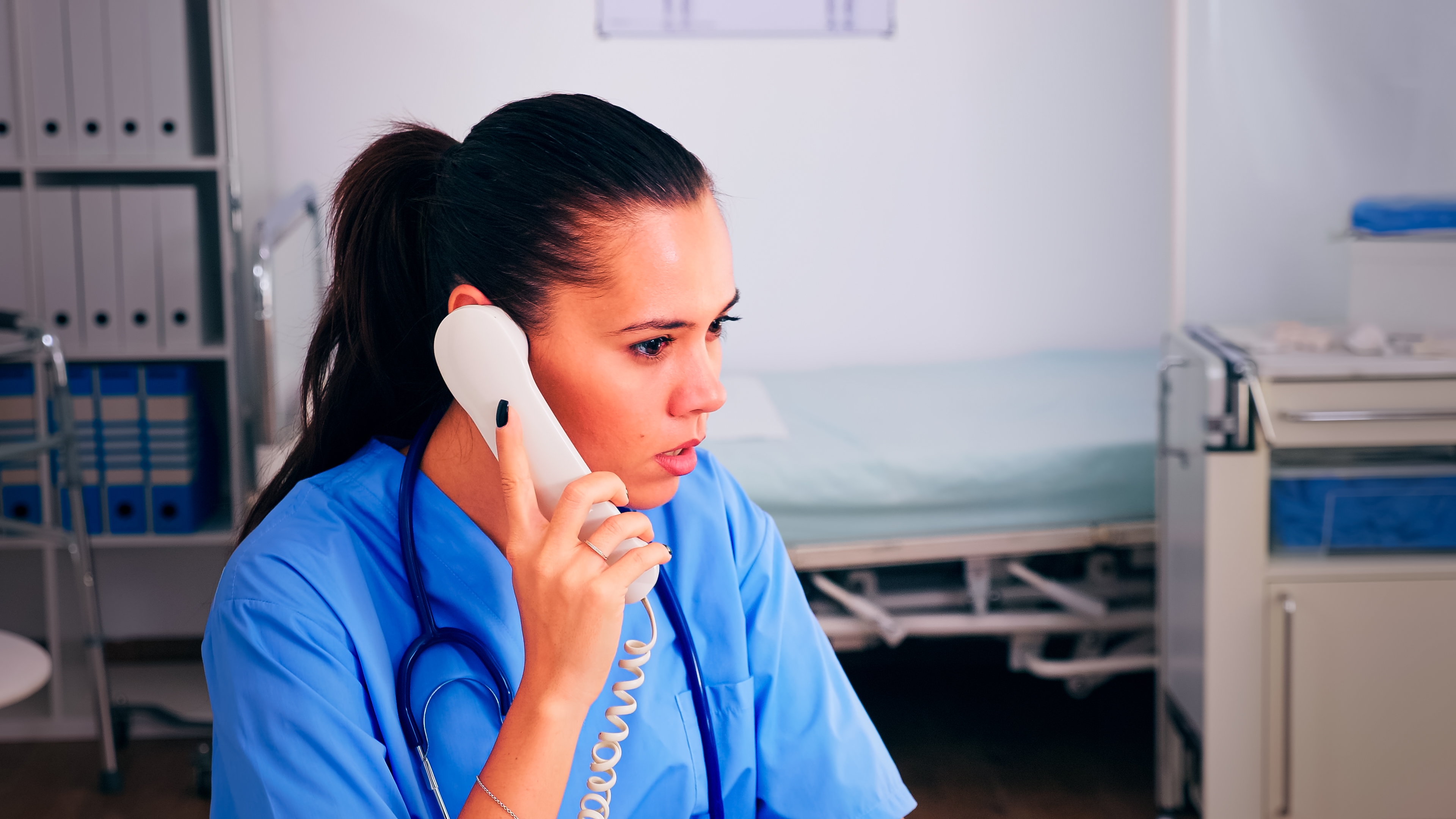A healthcare worker in scrubs is on the phone in a medical setting, appearing attentive. A hospital bed and medical equipment are visible