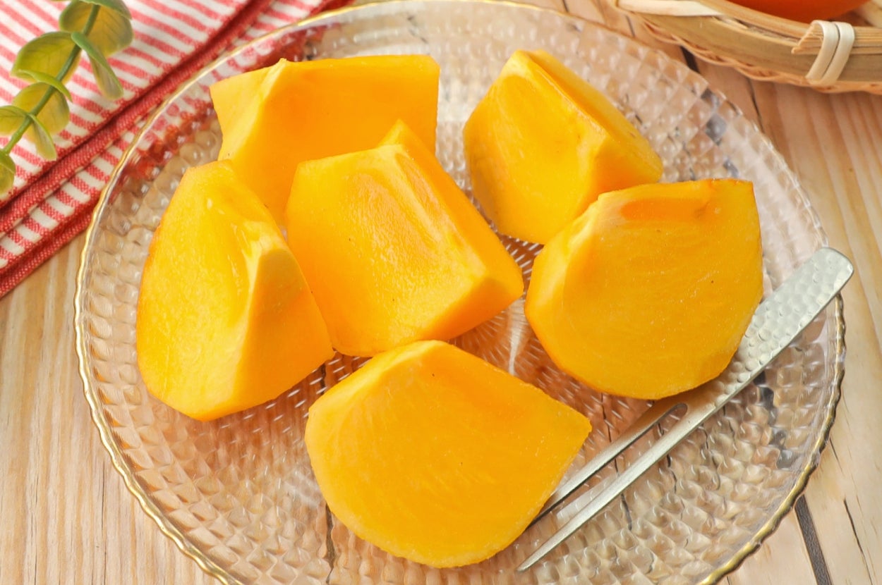 ガラスの皿にカットされた柿の実が盛られ、横にデザートフォークが置かれている。背景にバスケット入りの柿と布。