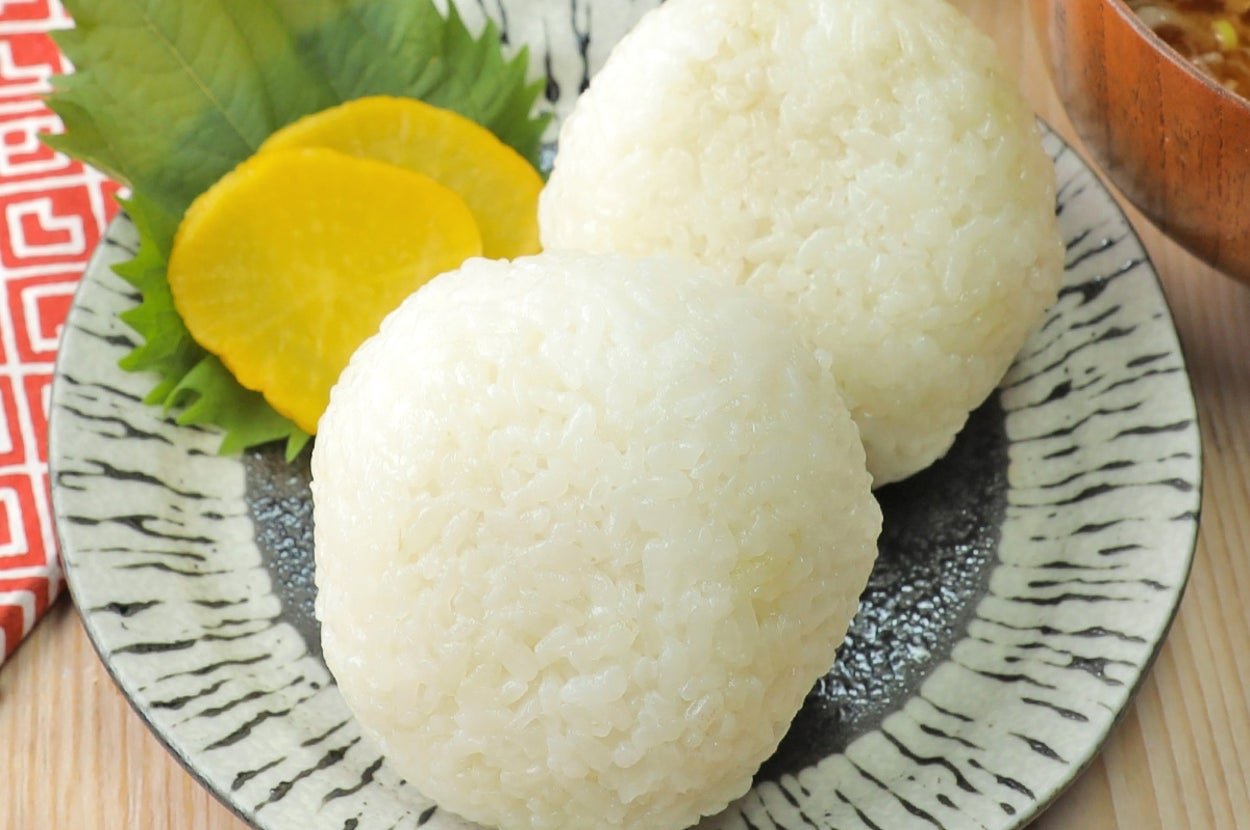 お皿に盛られた2つのおにぎりと、添えられたたくあん。背景には赤い布とみそ汁。