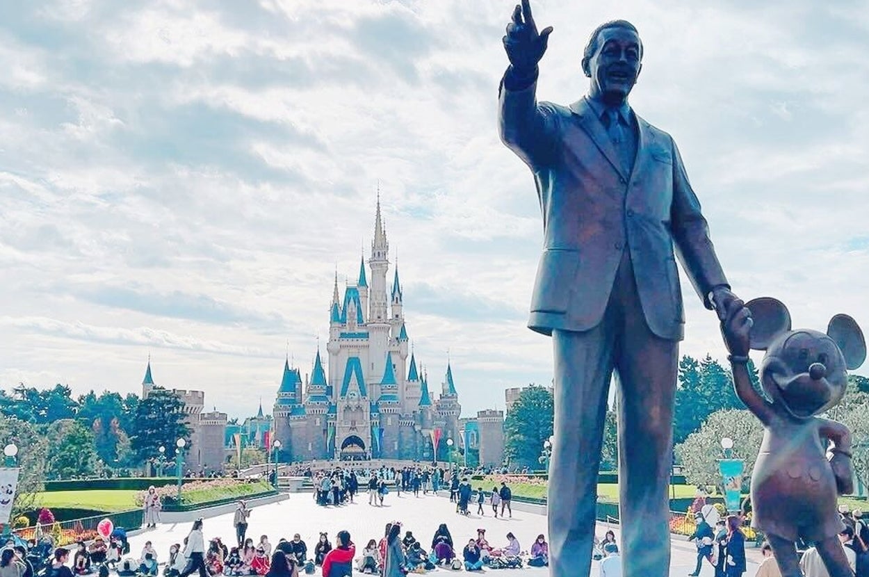 ディズニーキャラクターの像が建つテーマパークで、人々が楽しんでいる様子。背景には城。