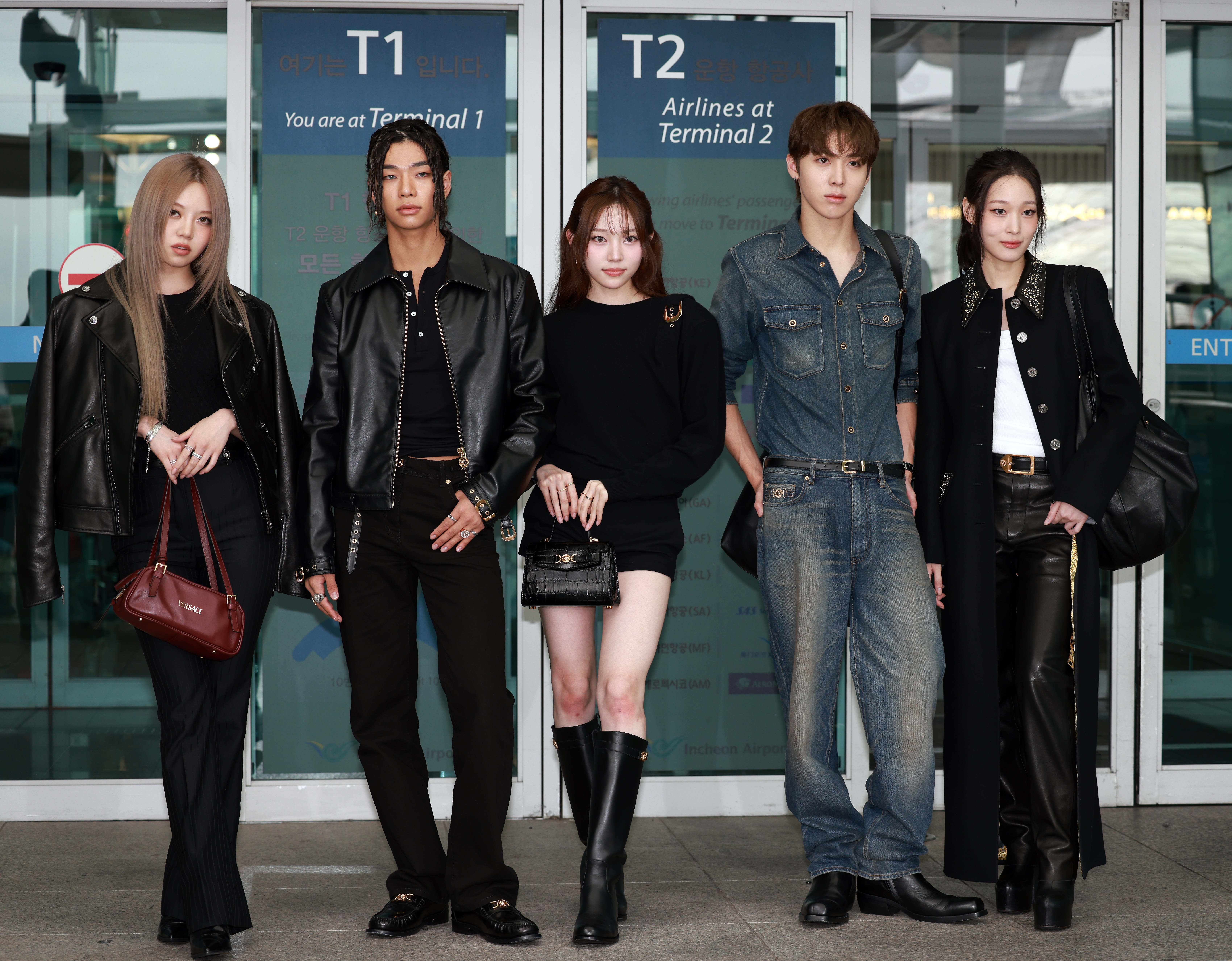 Five individuals stand in stylish outfits outside an airport terminal: three in black attire, one in denim, and the other in a leather jacket and pants