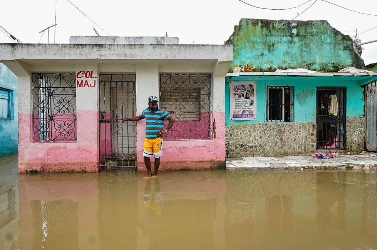 A person stands in knee-deep floodwater in front of a small, worn building with barred windows, highlighting flood conditions in a residential area