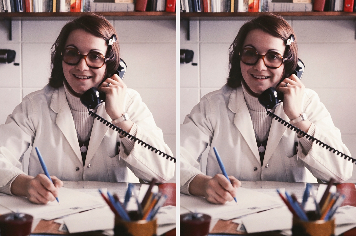 Person wearing glasses and a lab coat, talking on a corded phone while writing at a desk in an office with books on shelves