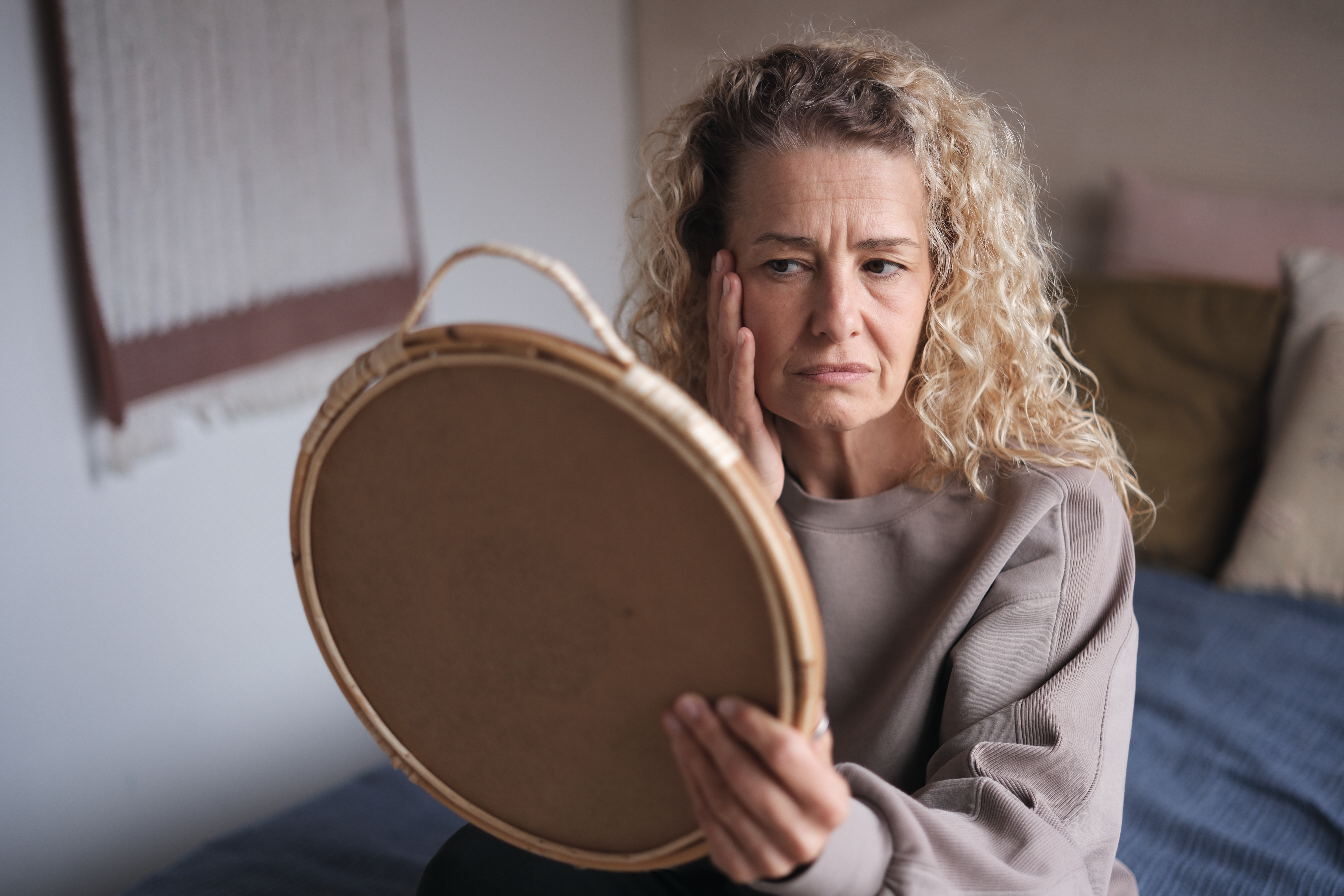 En kvinne med krøllete hår, iført en uformell genser, ser ettertenksomt inn i et rundt speil mens hun sitter på en seng