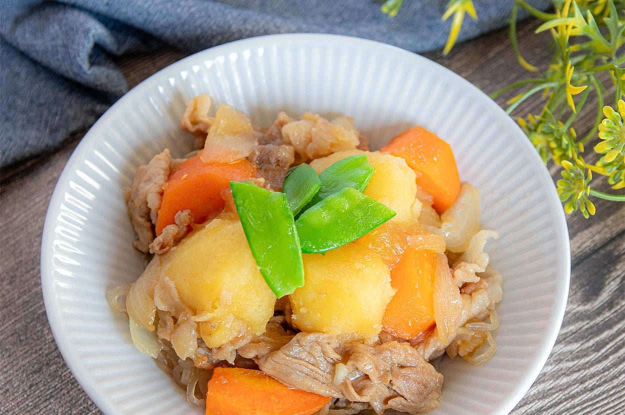 じゃがいも、にんじん、豚肉が入った煮物料理が白い皿に盛られています。テーブルには花とナプキンもあります。