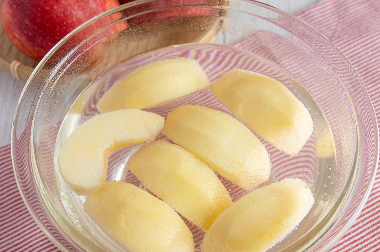 ボウルに浸されたリンゴのスライスとバックに見える丸ごとのリンゴ。テーブルには縦縞の布地が敷かれています。