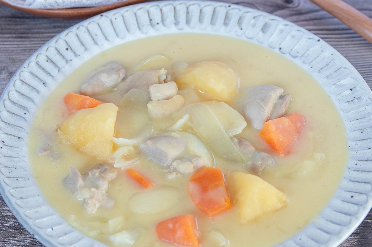 じゃがいも、　にんじん、鶏肉が入ったクリーミーなシチューの皿。背景にパンと木のスプーンがある。