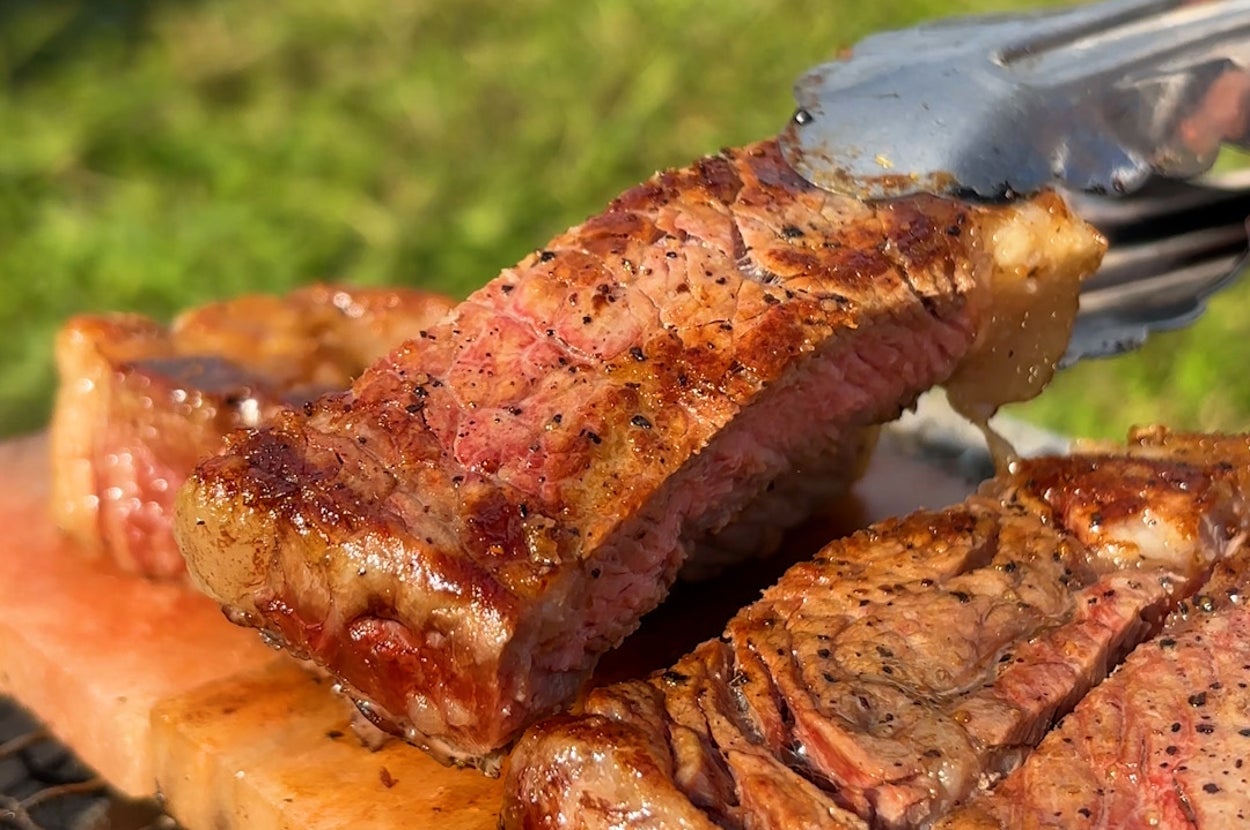 焼き網の上でジューシーに焼かれたステーキがトングで持ち上げられている。背景に芝生。