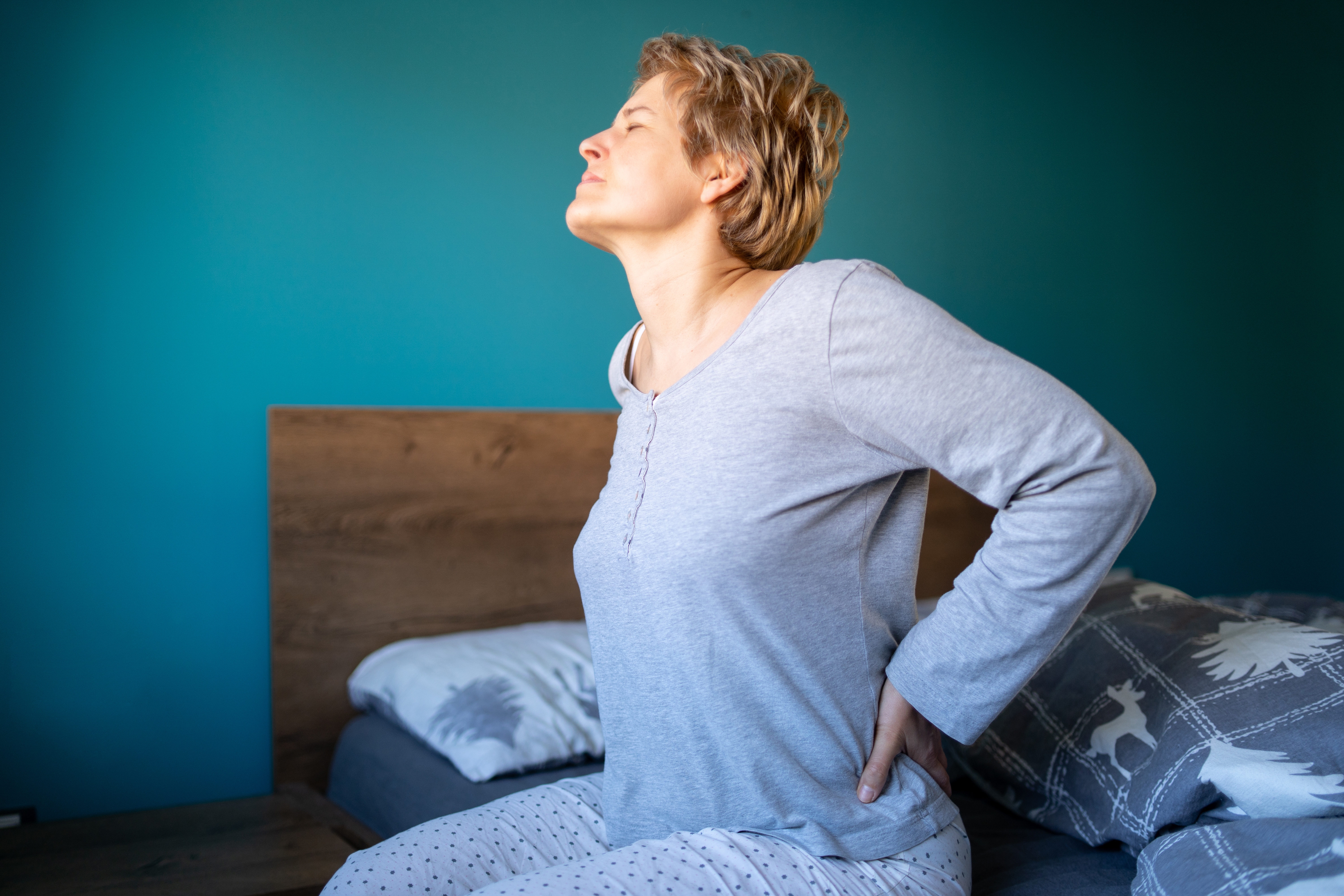 Person i pyjamas som sitter på en seng, strekker seg med hendene på korsryggen, opplever muligens ubehag. Rommet har en enkel, koselig følelse