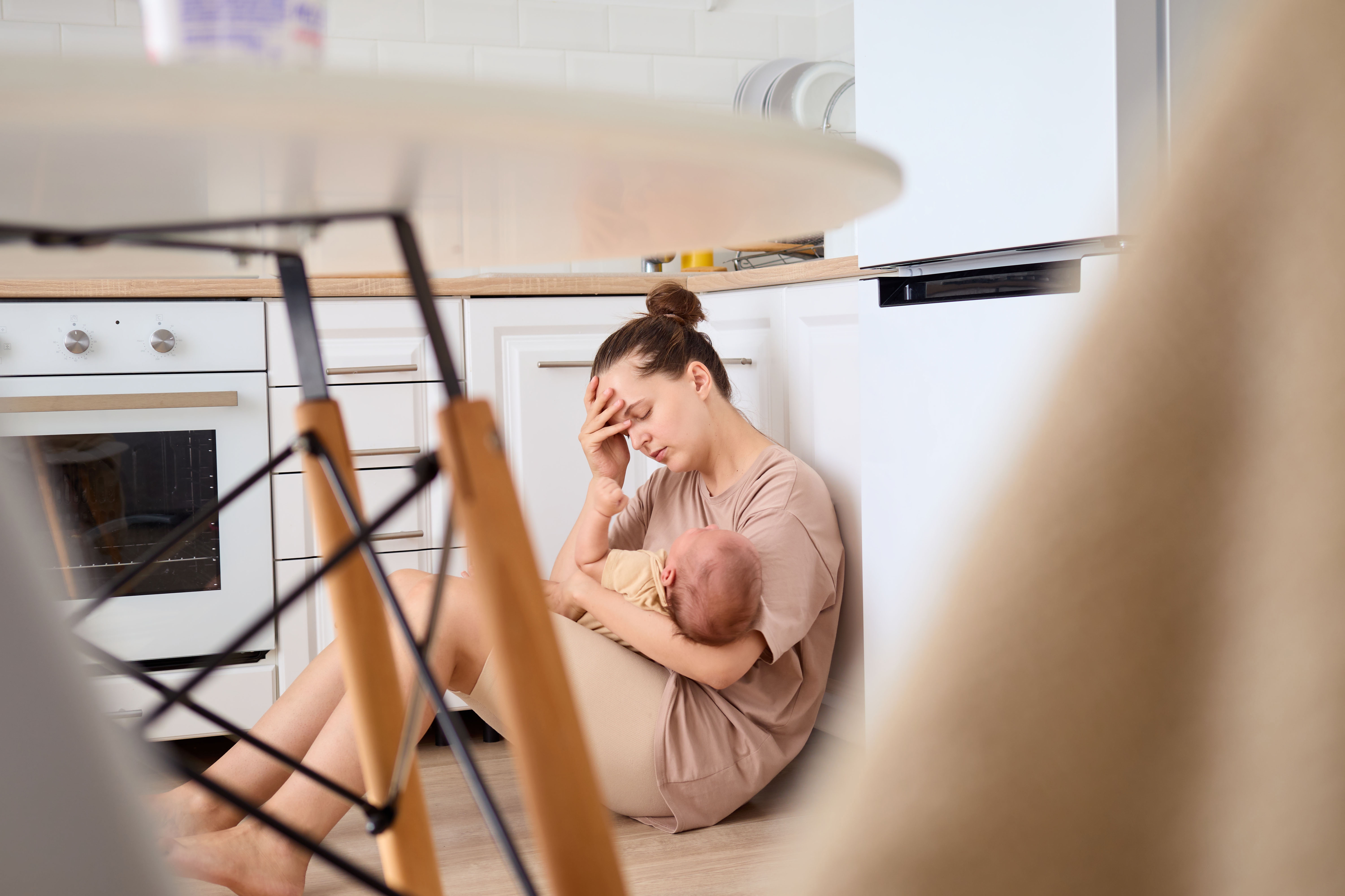 En sliten kvinne sitter på et kjøkkengulv og vugger en sovende baby mens hun hviler hodet i hånden, formidler utmattelse og ømhet