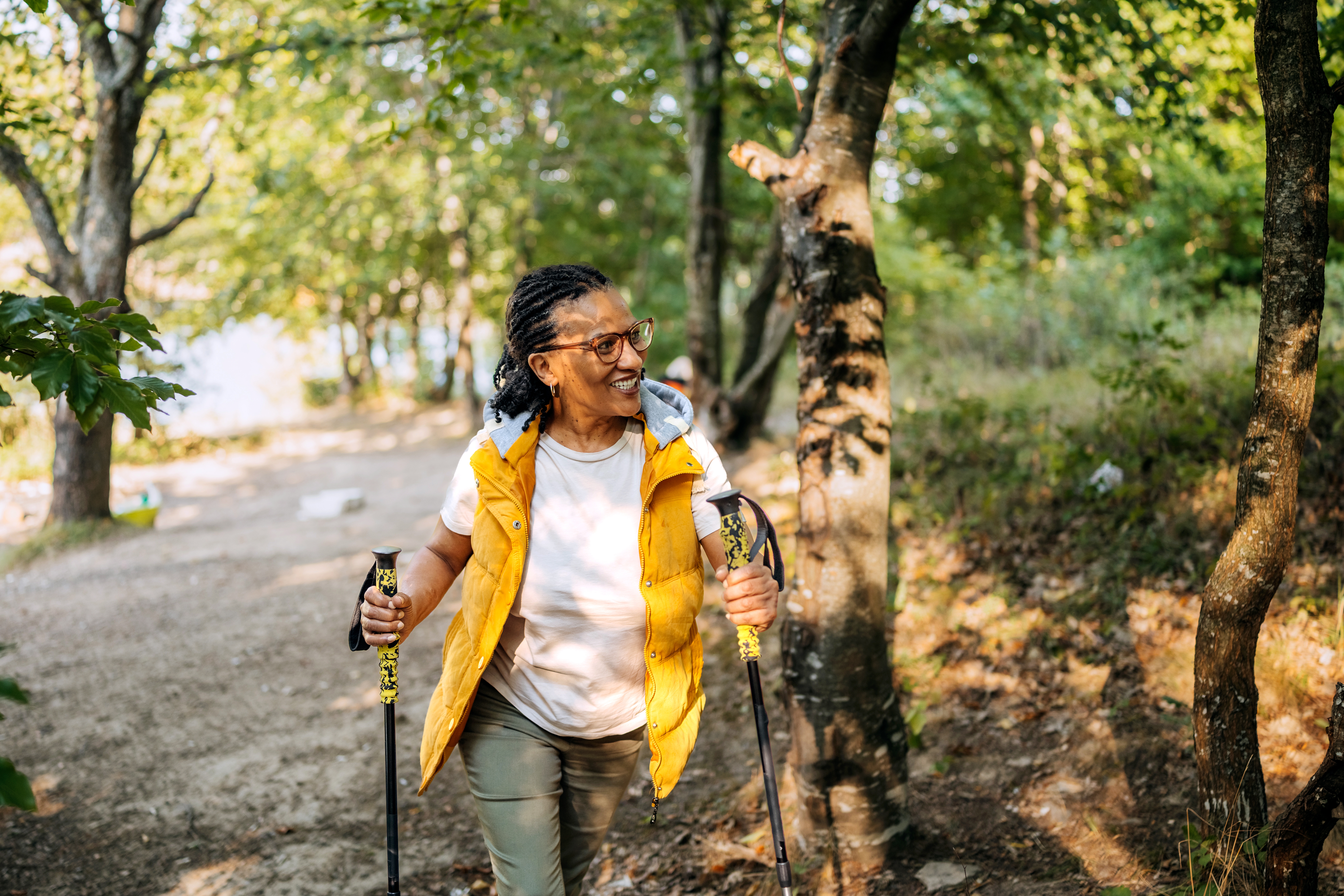 Person som smiler mens han går tur med staver på en skogssti, iført en oppblåst vest og t-skjorte. Sollyset filtrerer gjennom trærne og skaper en rolig atmosfære