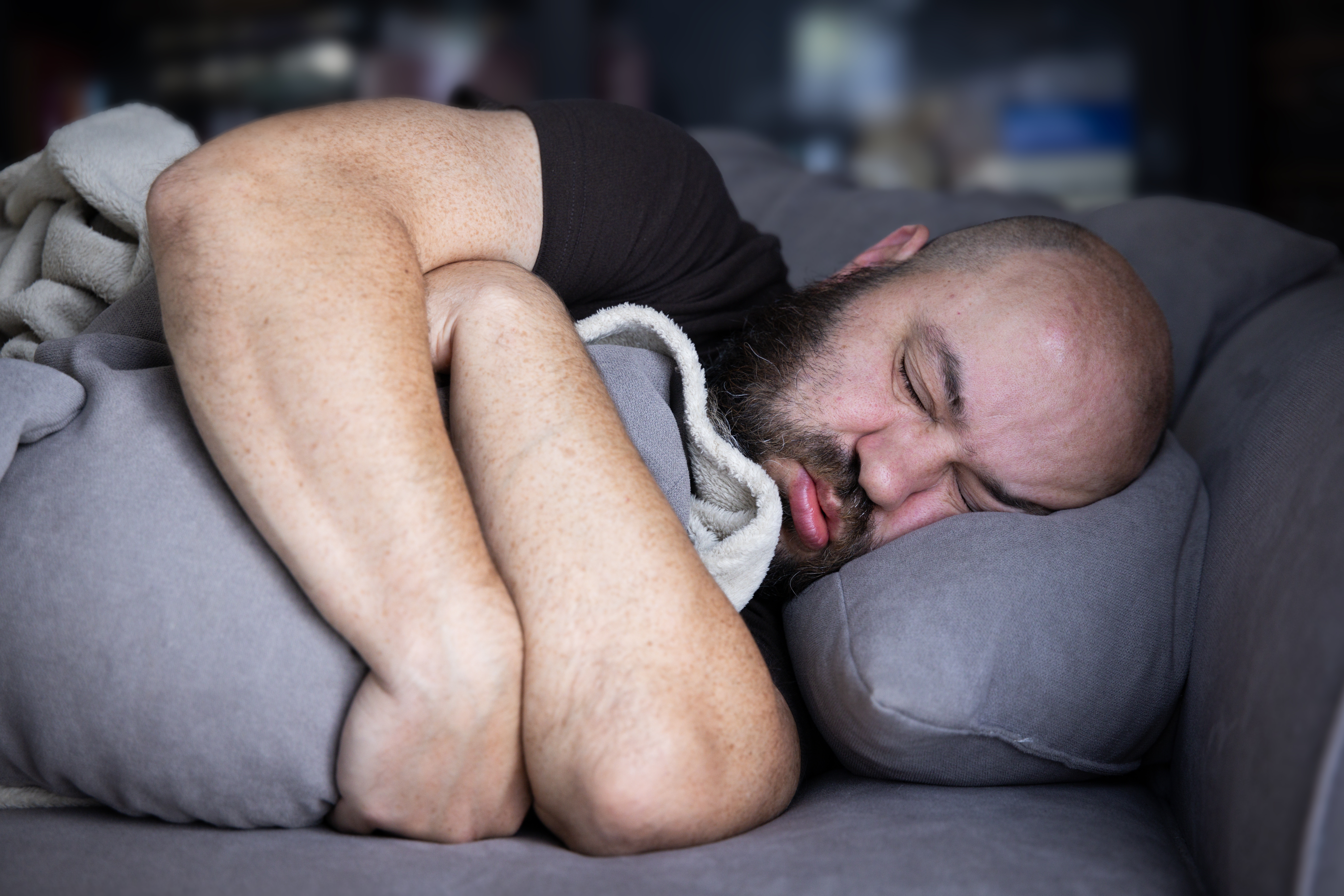En person med skjegg sover på en sofa, pakket inn i et teppe, med et fredelig uttrykk