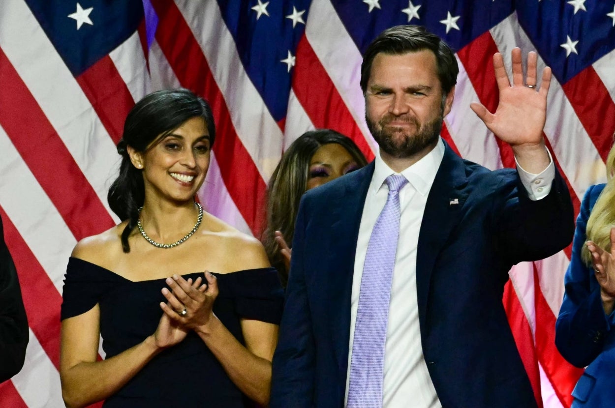 A man in a suit waves on stage, standing next to a woman in an elegant off-shoulder dress, with U.S. flags in the background
