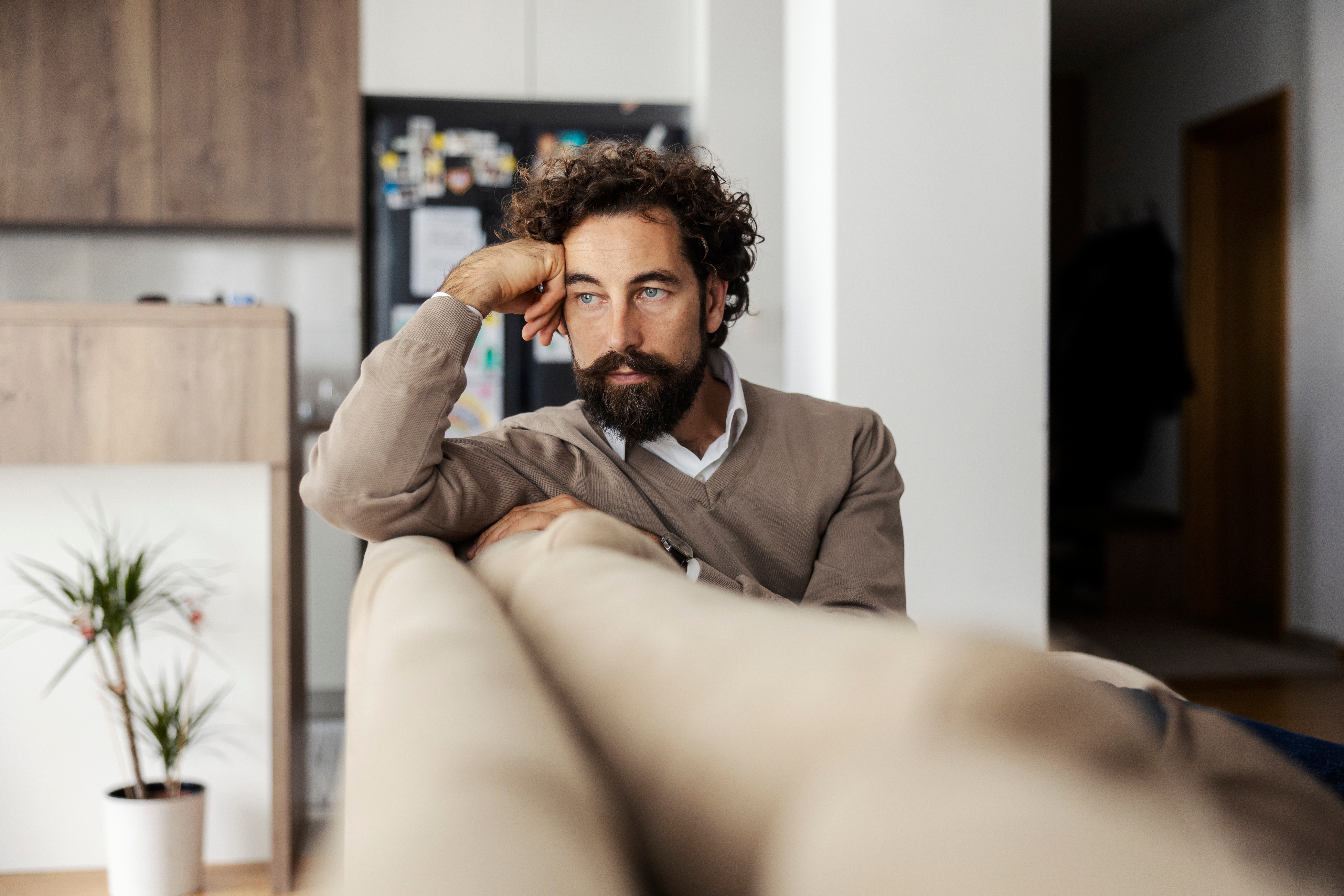 En person med krøllete hår og skjegg, iført en uformell genser, lener seg omtenksomt på en sofa i en moderne stue