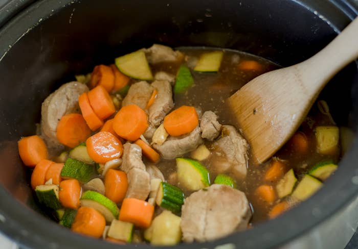 A slow cooker with a hearty stew of sliced carrots, zucchini, and chunks of meat being stirred with a wooden spoon