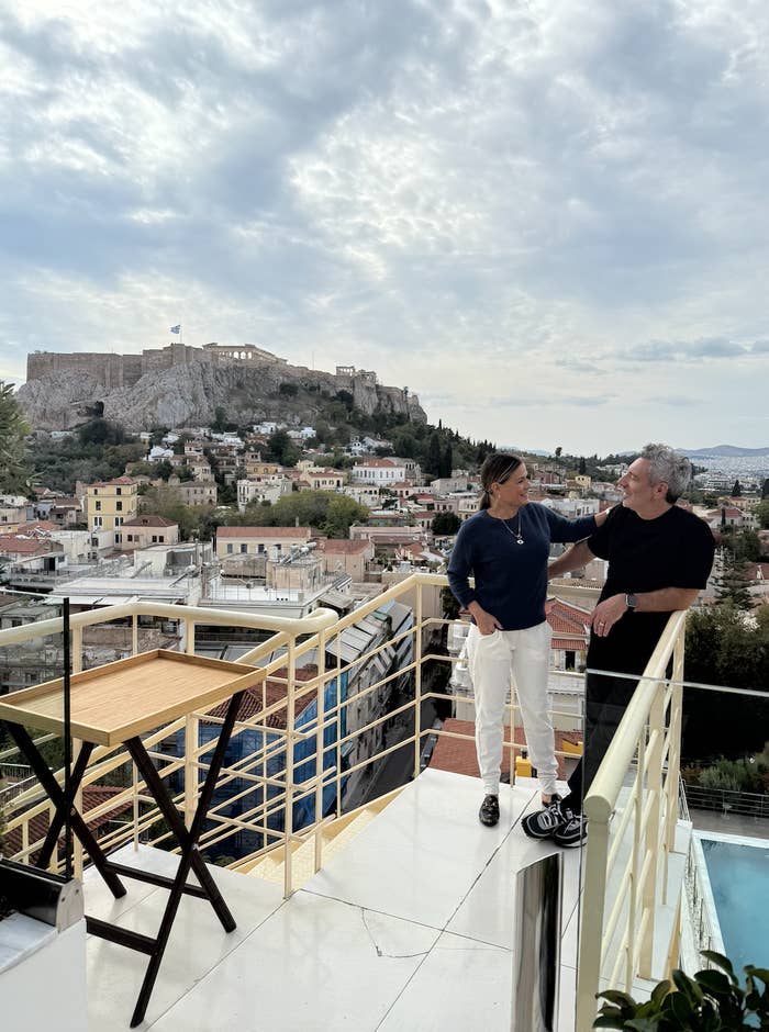 Two people stand on a balcony overlooking Athens, with the Acropolis in the background. They appear engaged in conversation