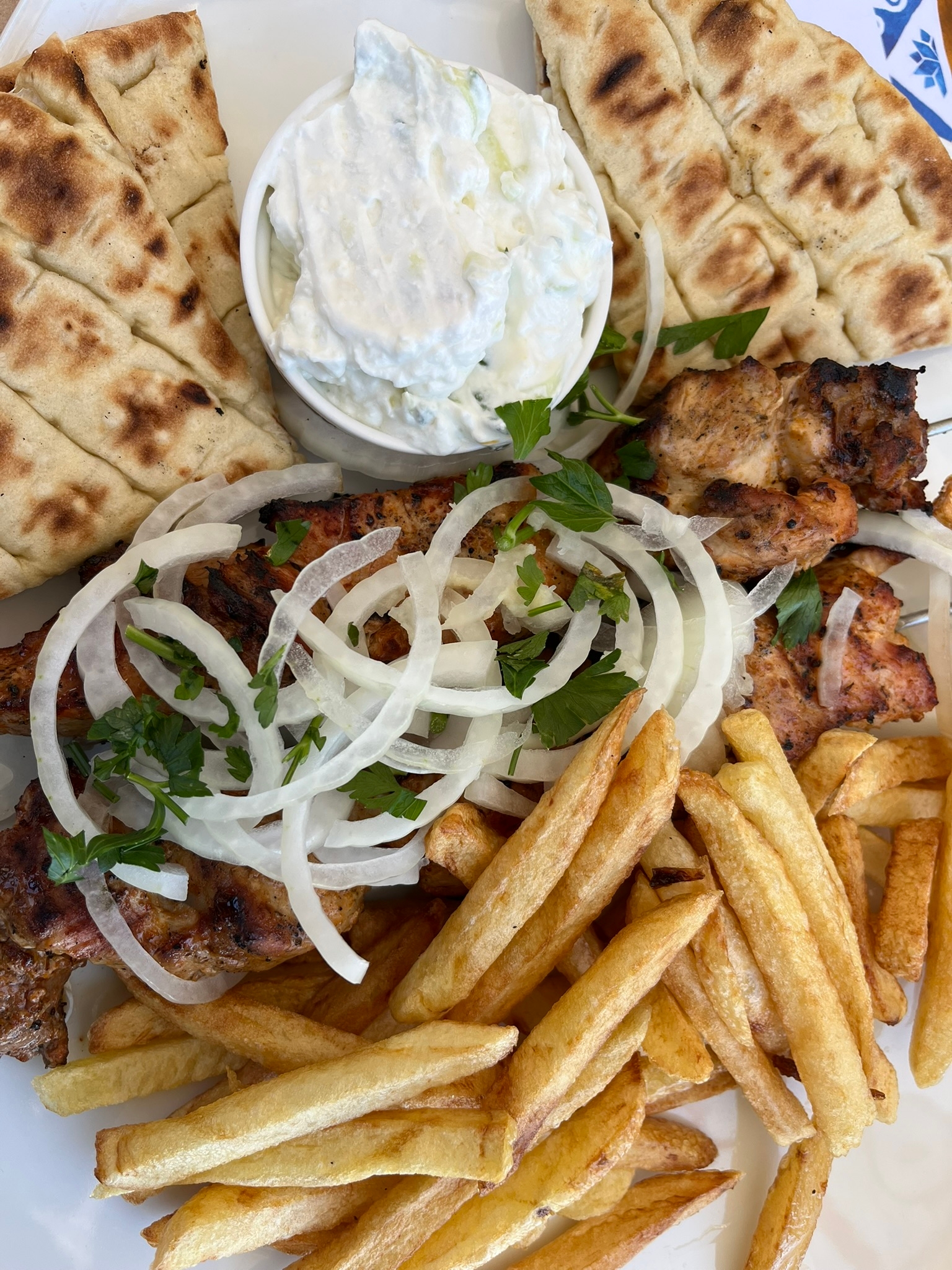 Plate of grilled chicken souvlaki with fries, sliced onions, pita bread, and tzatziki sauce garnished with parsley