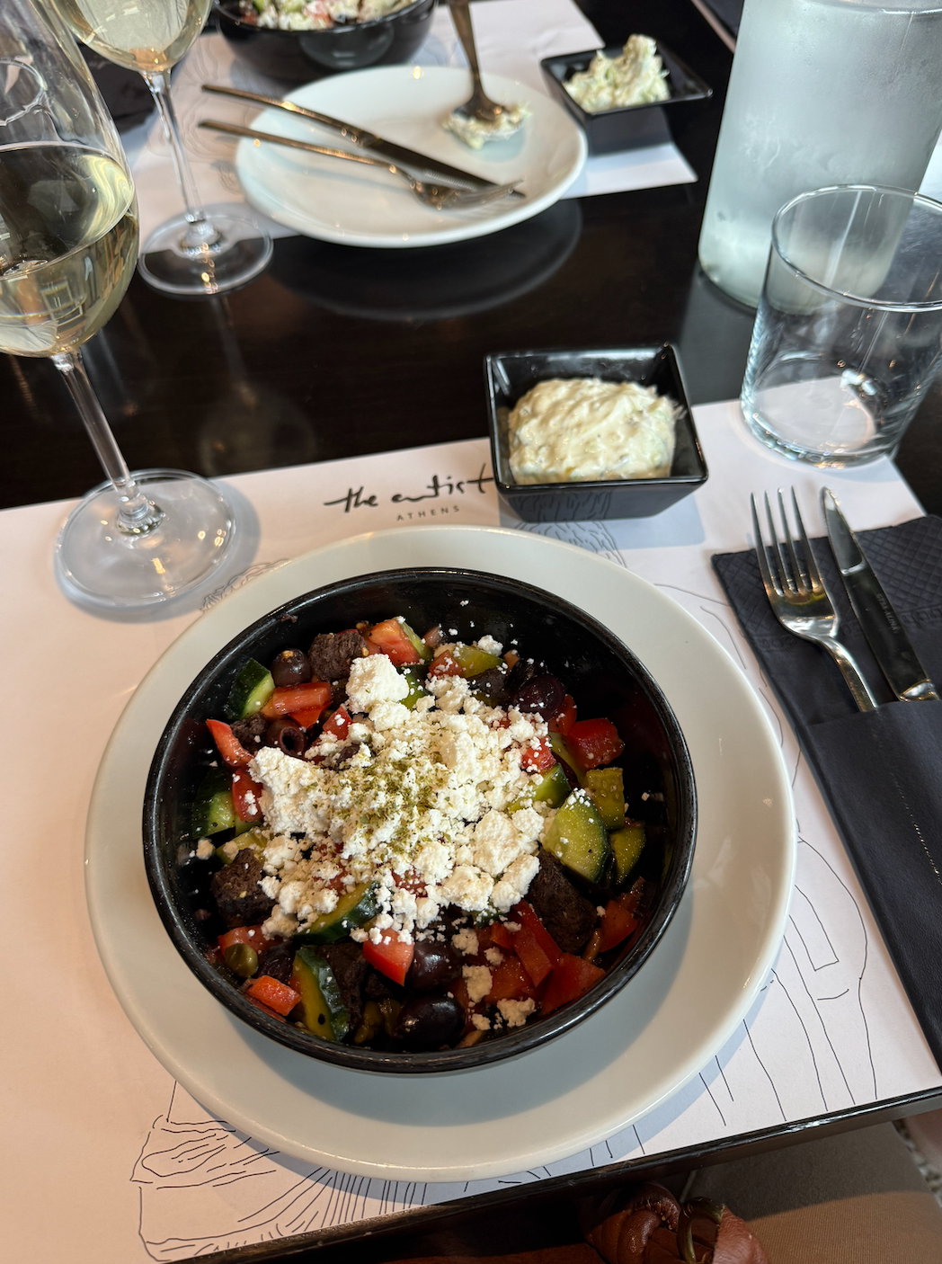 Bowl of Greek salad with feta, olives, peppers, and cucumbers on a restaurant table set with wine glasses and cutlery