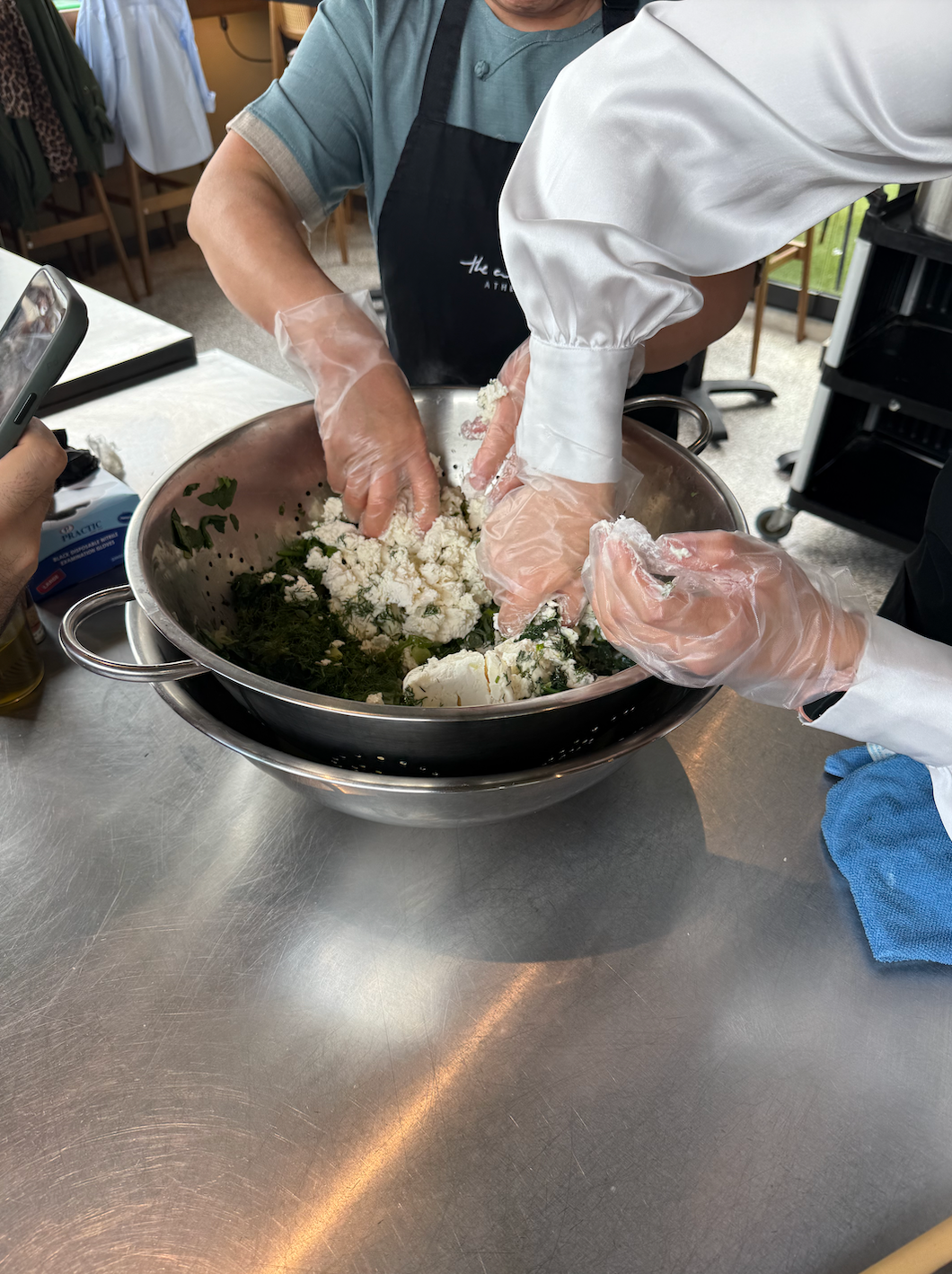 Two people wearing gloves mix ingredients and feta in a large bowl on a kitchen counter