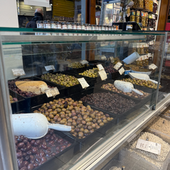 An assortment of olives and beans displayed in trays at a market counter, with scoops for serving. Various signs are visible above the products