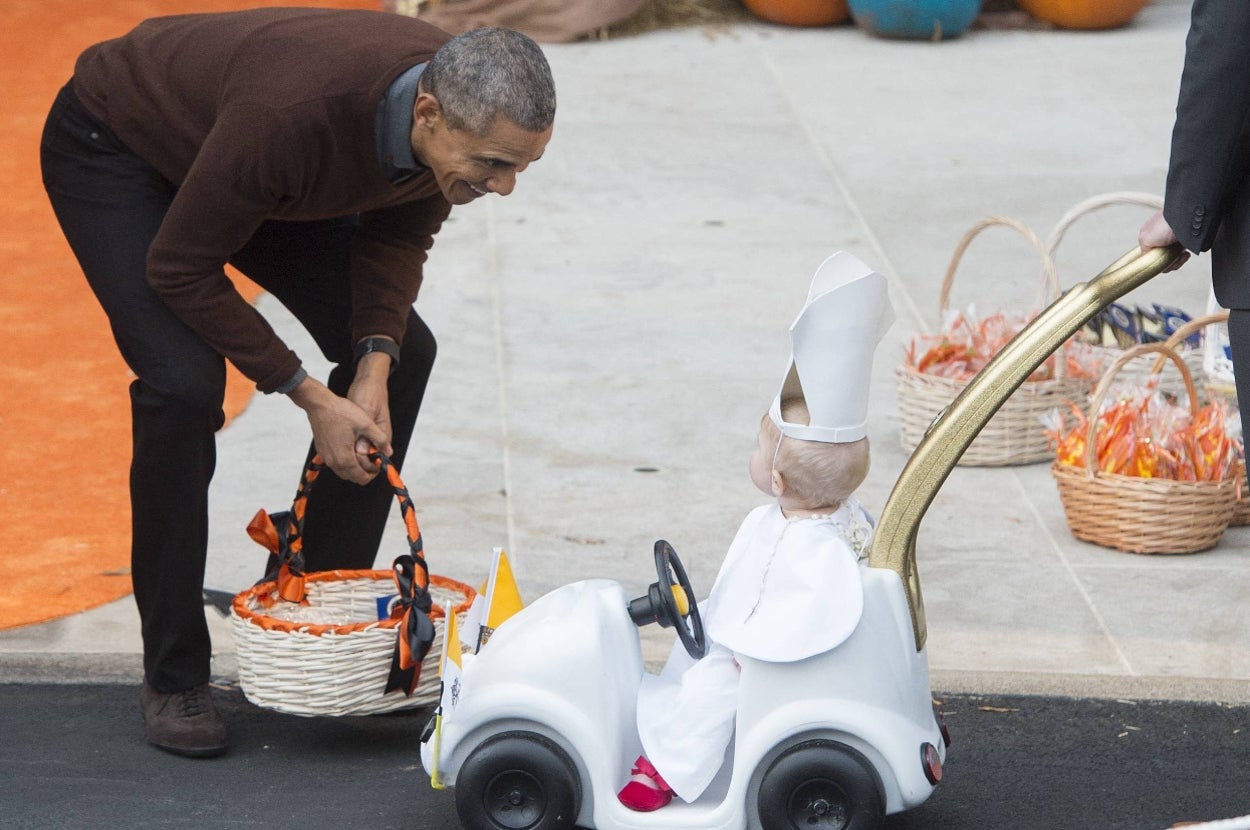 A man smiles at a toddler dressed as a Pope in a costume car during a Halloween event, with baskets of candy in the background