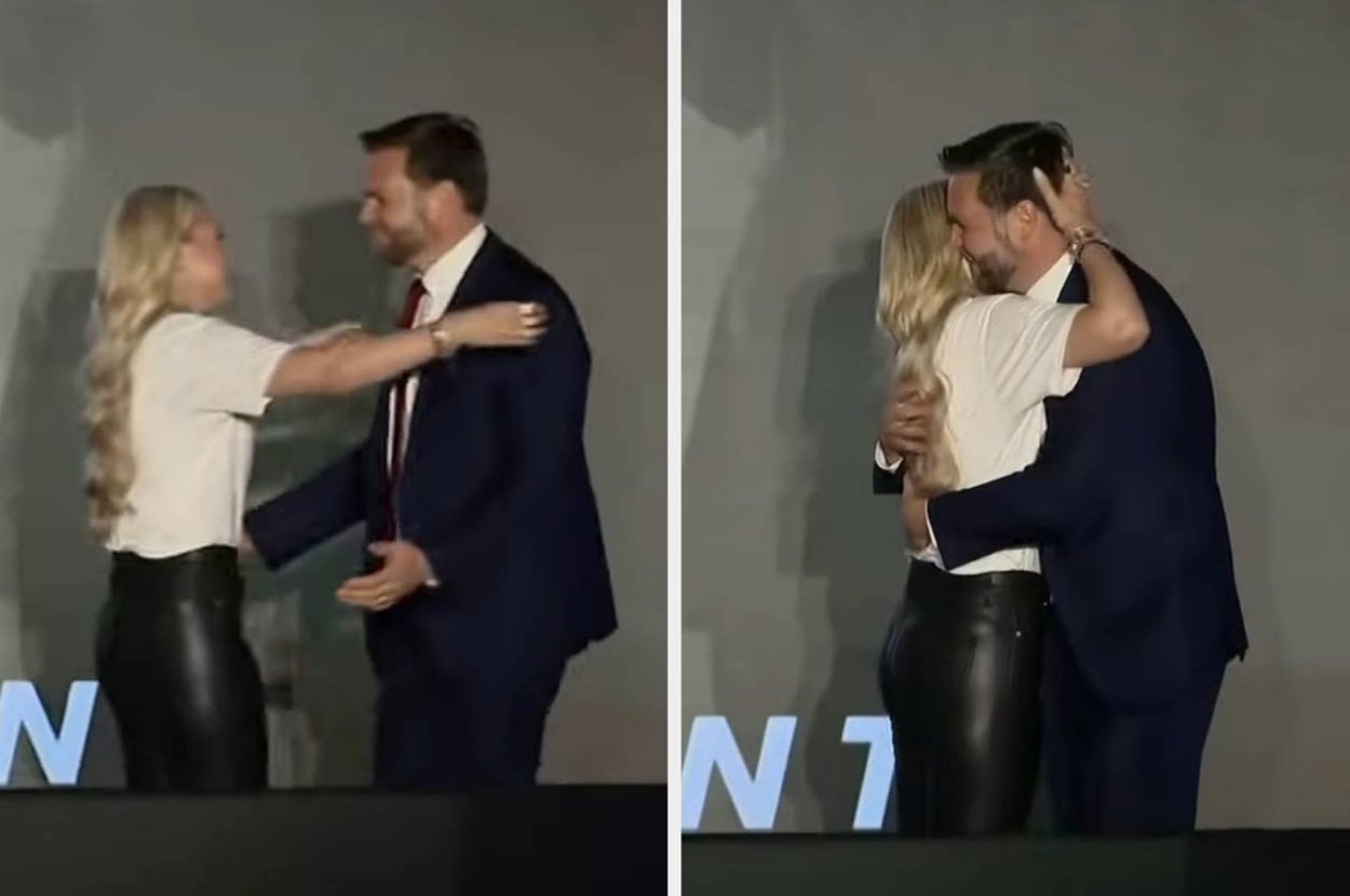Two people embrace on stage at an event. One wears a suit, the other is in a white shirt and leather pants