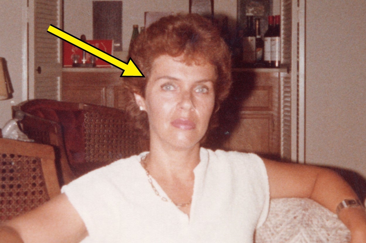 Person sitting indoors, pointed to by an arrow, wearing a light short-sleeve top, and a necklace, in a casual setting