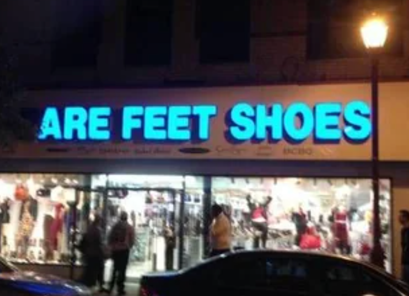Storefront with a motion   speechmaking  "ARE FEET SHOES" supra  a lit-up model   display. People and cars are disposable   outside