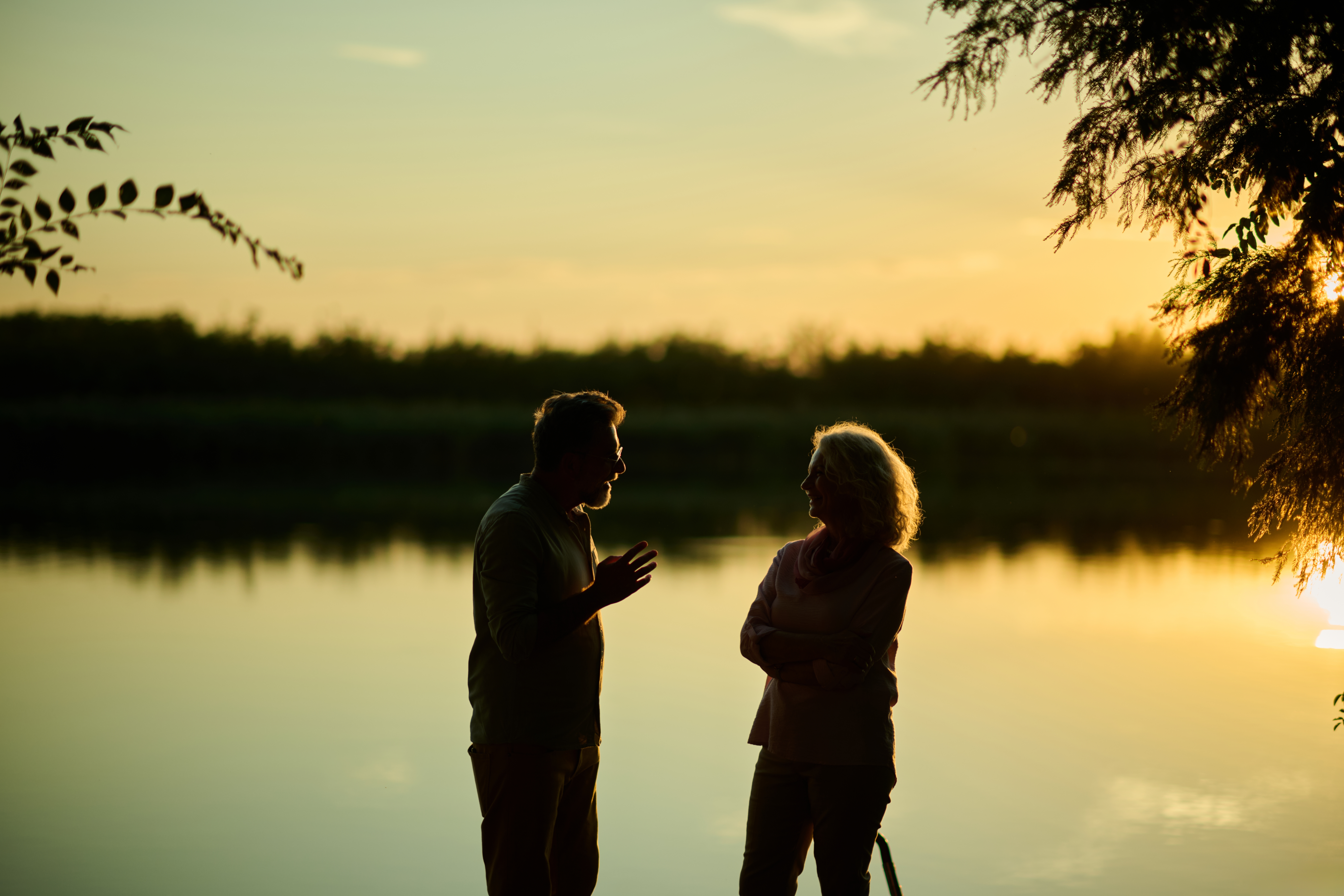 To personer er silhuettert mot en solnedgang ved en innsjø, engasjert i samtale, omgitt av trær, og skaper en fredelig utendørsscene