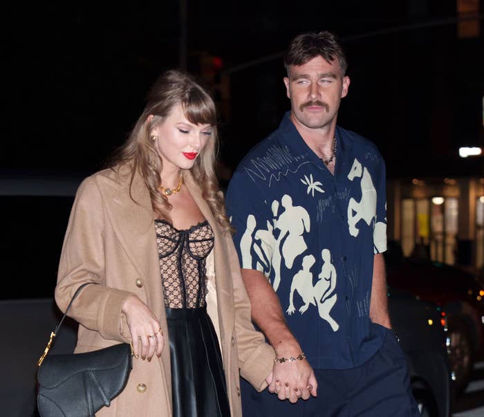 Taylor Swift in a lace top and skirt and Travis Kelce in a patterned shirt, holding hands outside