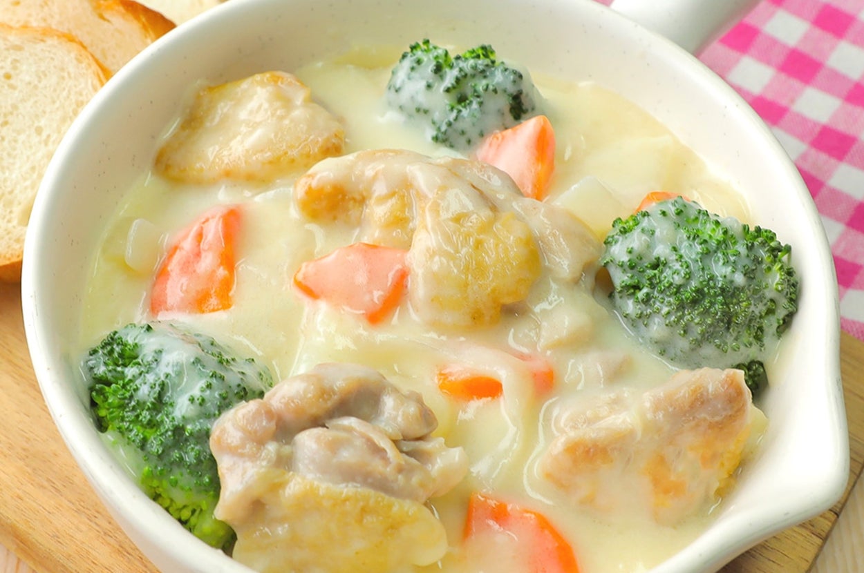 クリーミーなシチューに鶏肉、ブロッコリー、にんじんが入った料理が白い器に盛られている。背景にパンが置かれている。