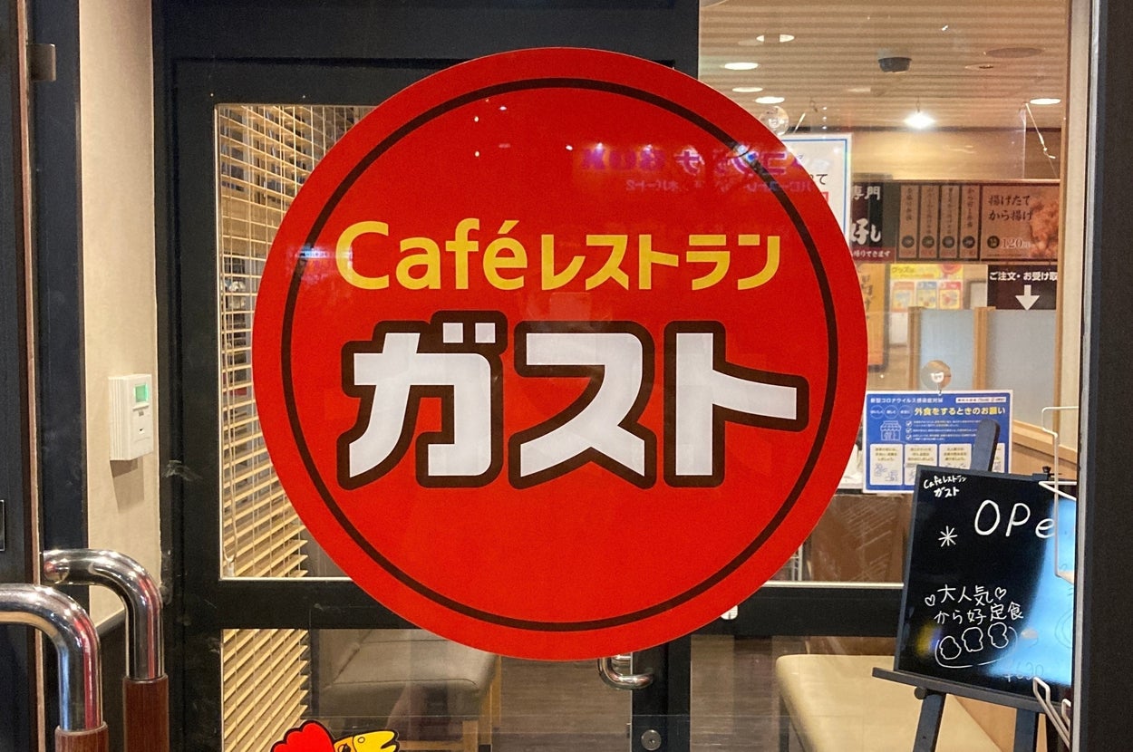 ファミリーレストラン「ガスト」の入り口ドア。営業案内が書かれており、店内が少し見える。