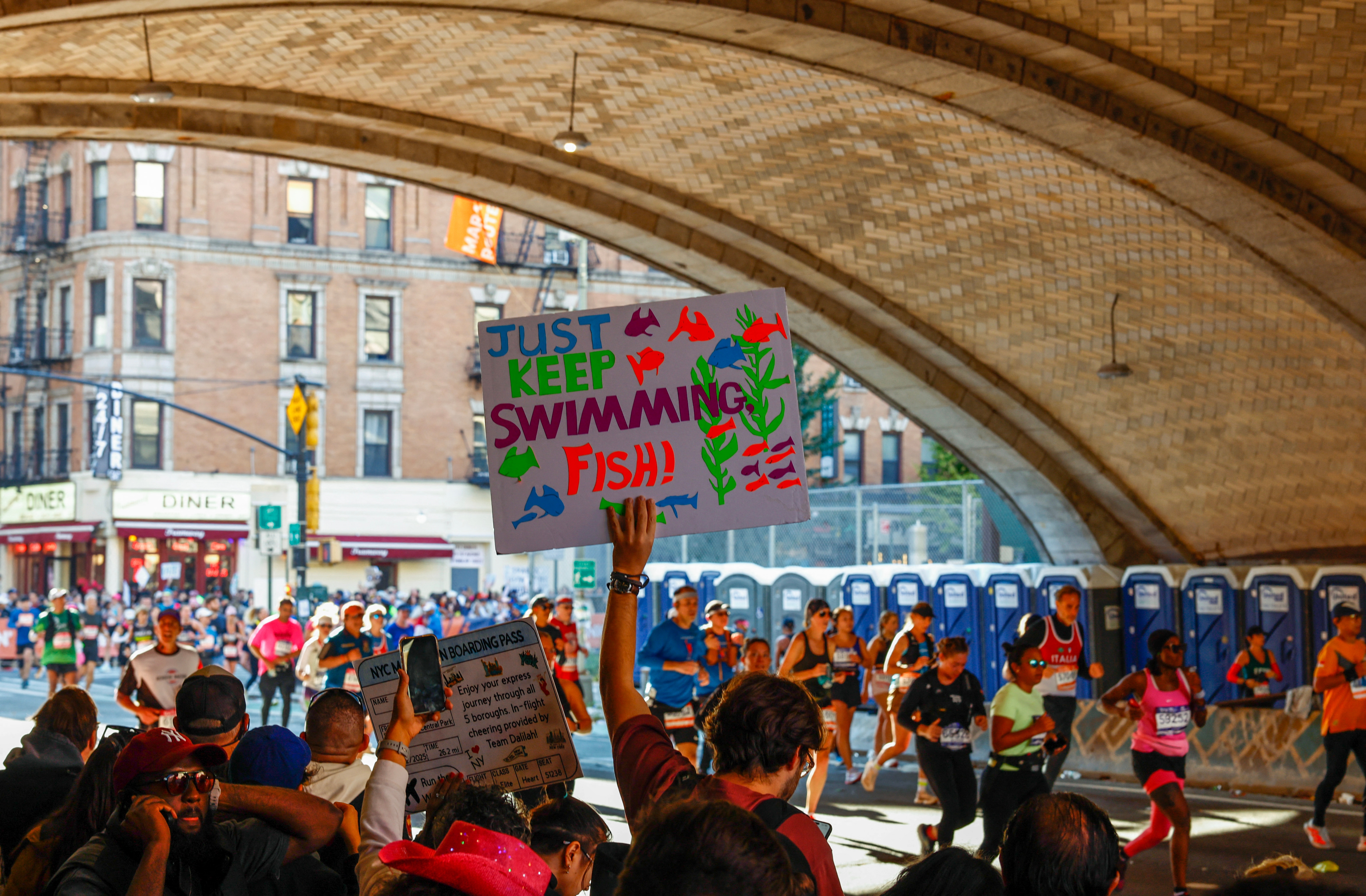 Person holding a colorful sign with "Just Keep Swimming Fish!" at an outdoor event with a crowd and runners passing by